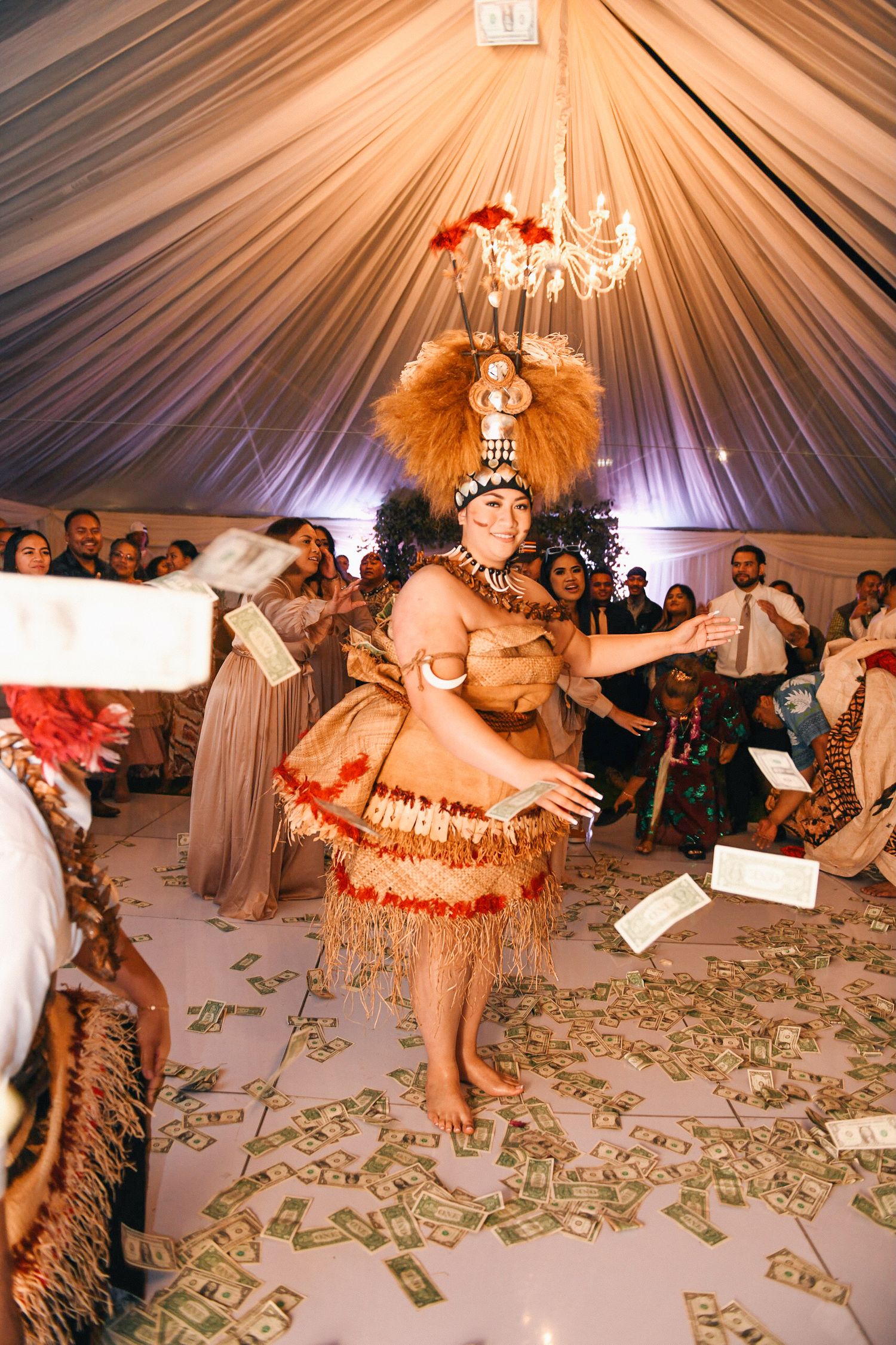 Polynesian dancer performs traditional dance in festive wedding reception tent with money on floor.