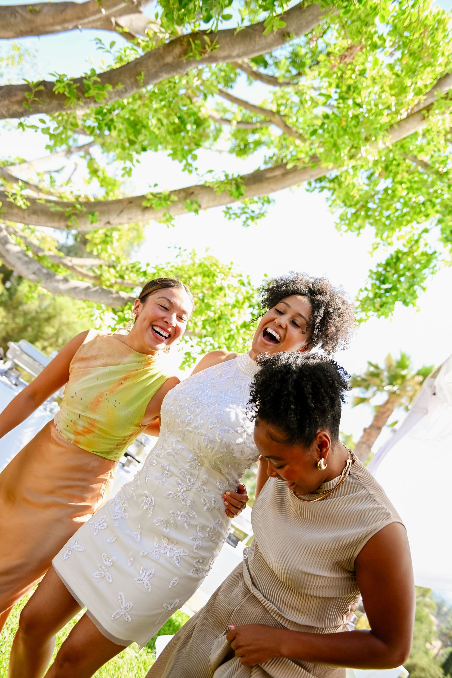 Three friends laughing and having fun together outdoors under green trees on a sunny day.