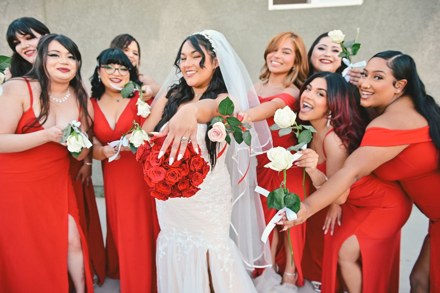 Bridesmaids in matching red dresses gather around bride in white gown showing off wedding rings at celebration.