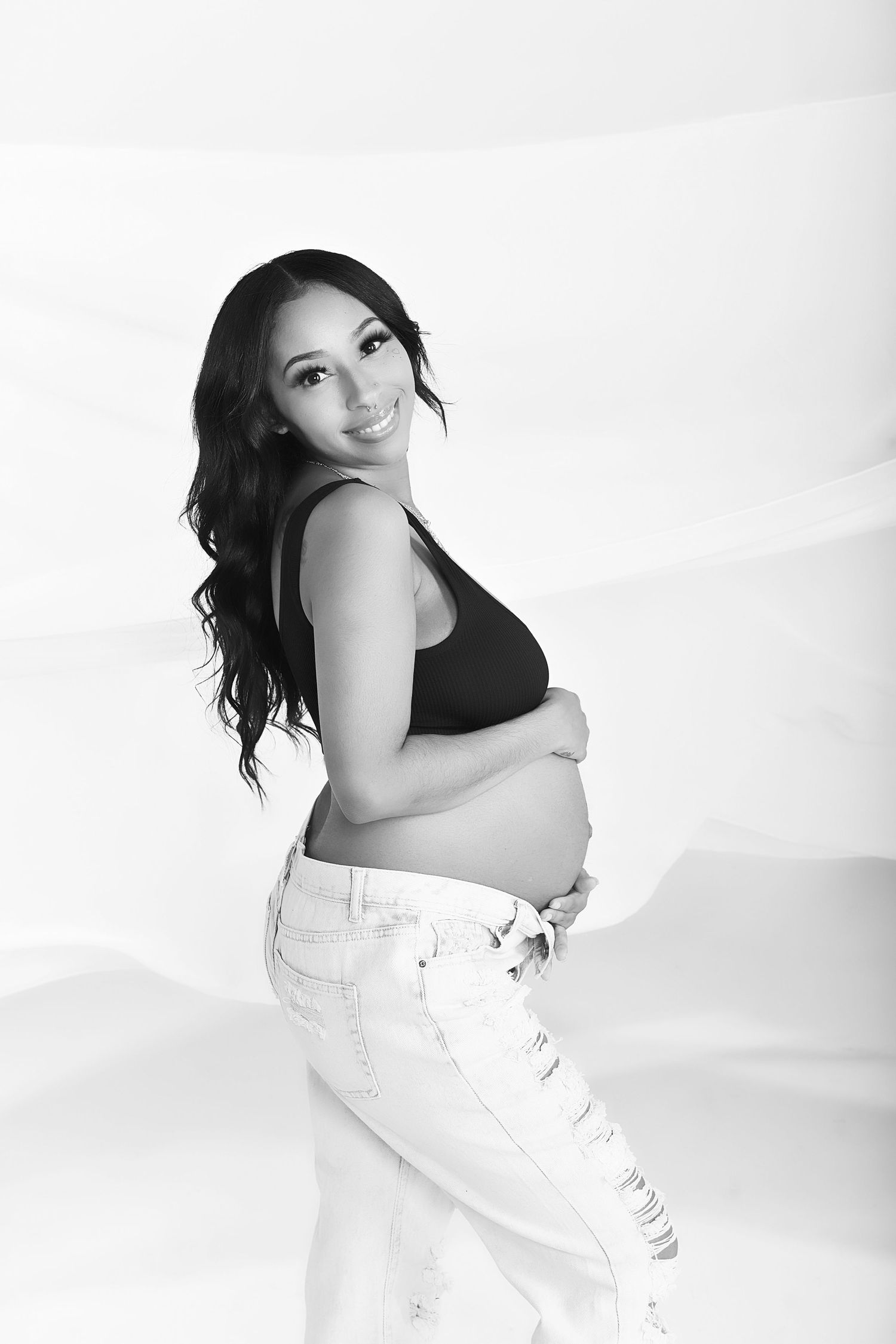 Black and white maternity photo showing an expectant mother in casual attire against a white background.