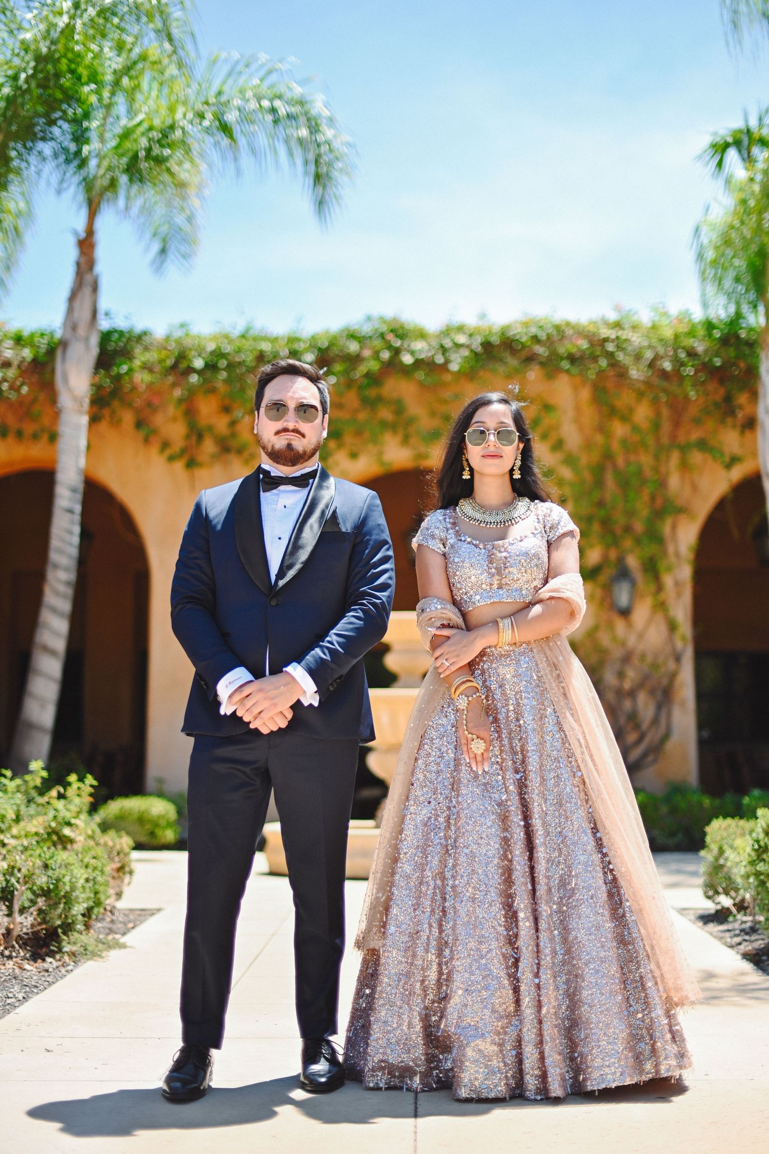 Couple in formal attire pose against Mediterranean style backdrop with palm trees.