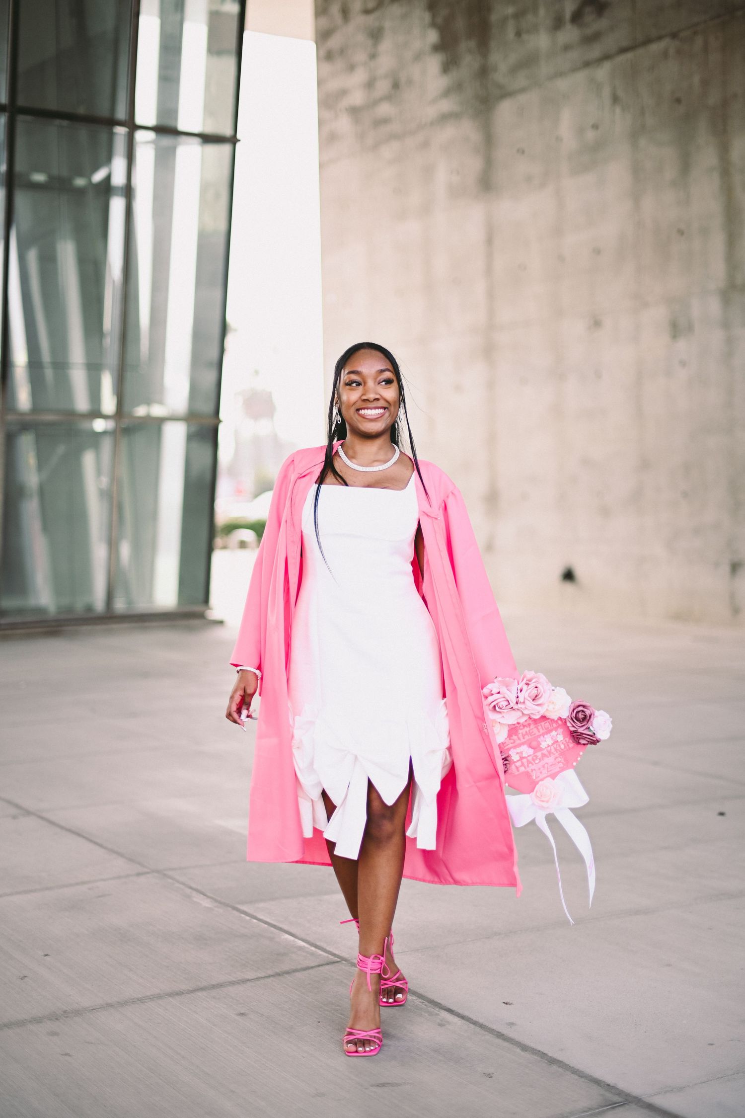 Stylish portrait of person in pink coat and white dress carrying flowers on urban sidewalk.