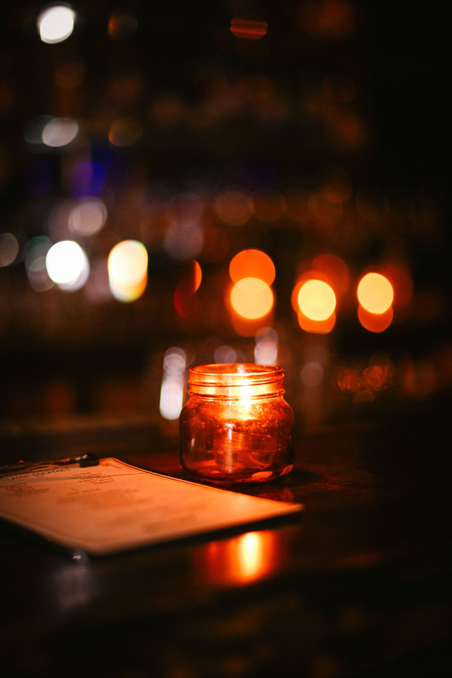 A glowing candle in a mason jar creates a warm ambiance against a dark background with bokeh lights.
