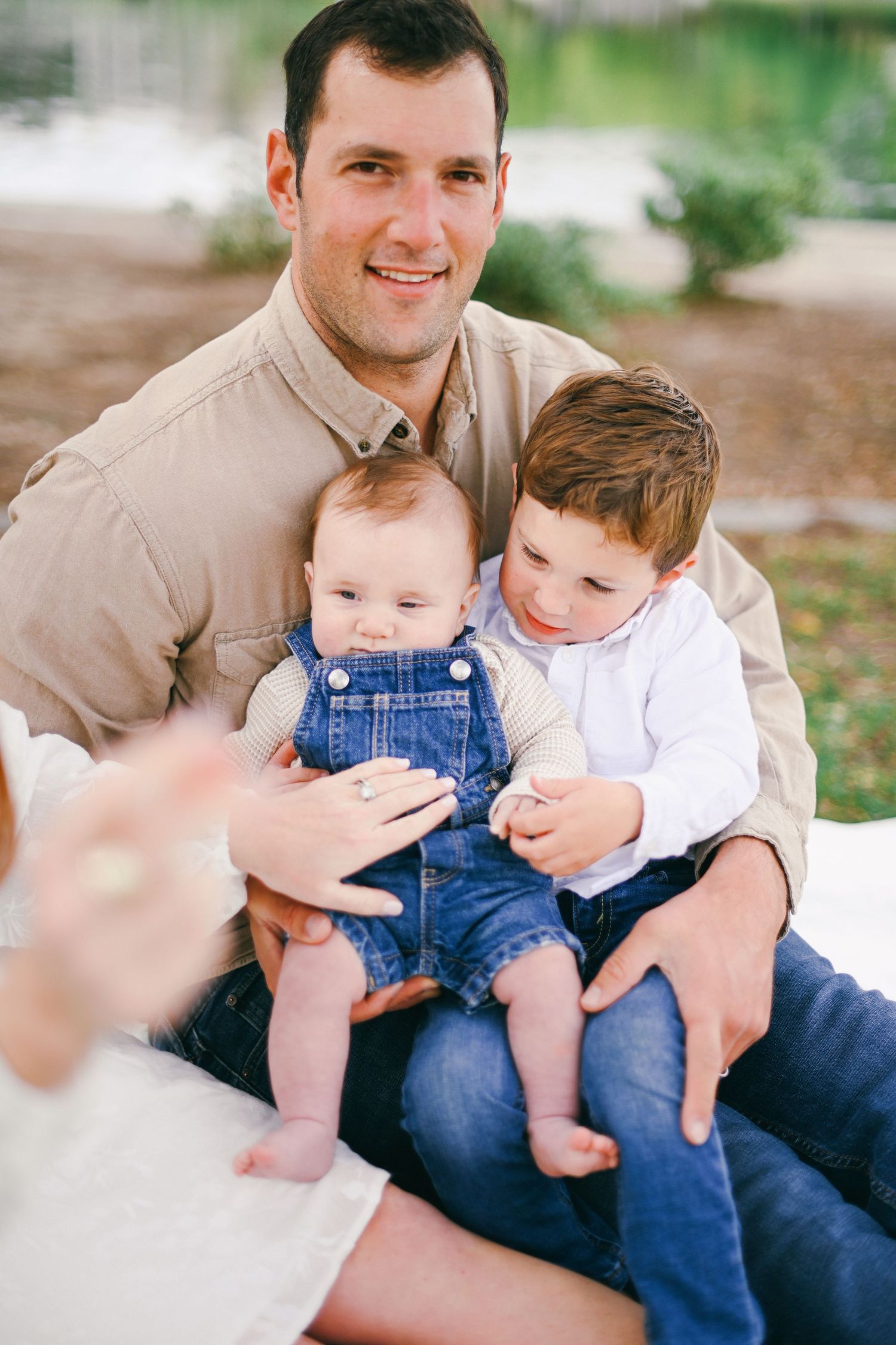 Father sits outdoors holding two young children in casual family portrait.