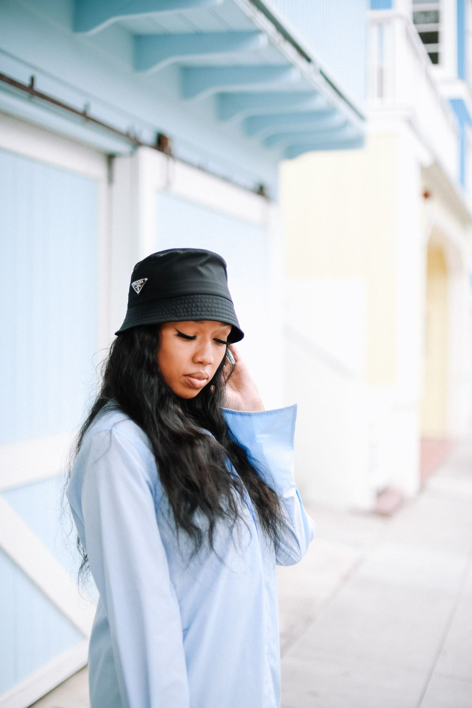Person in bucket hat and oversized shirt posing against blue wall.