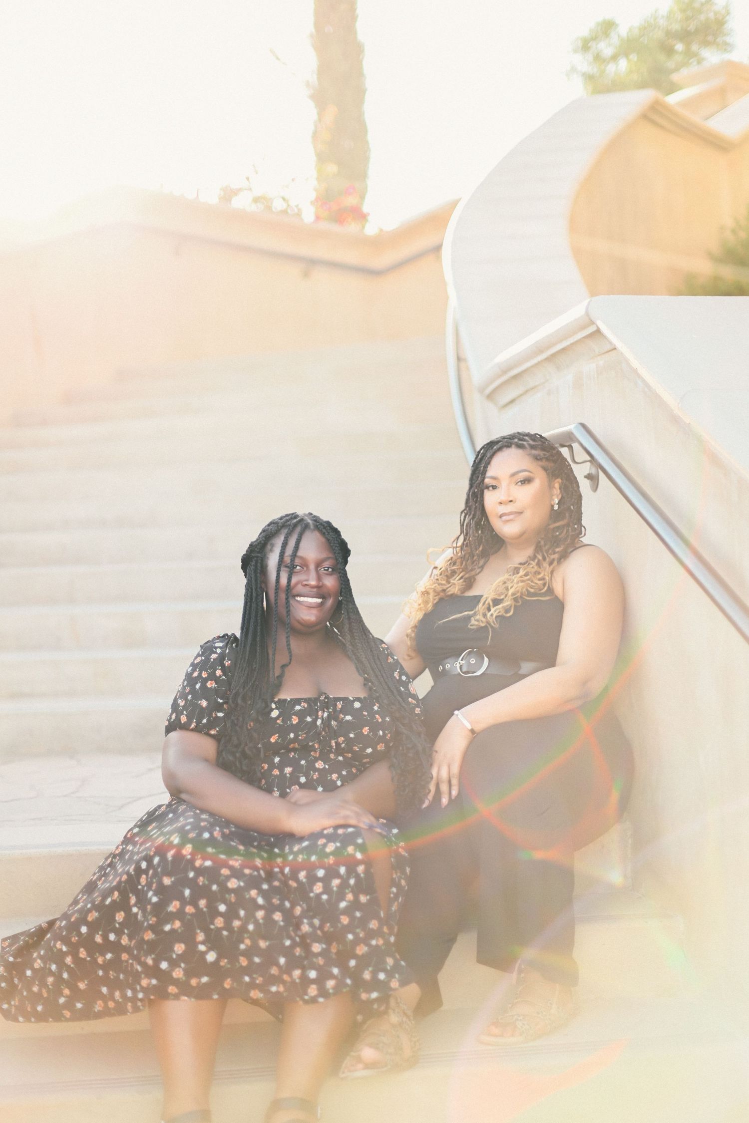 Two women in polka dot dresses pose together in golden sunlight near stairs.