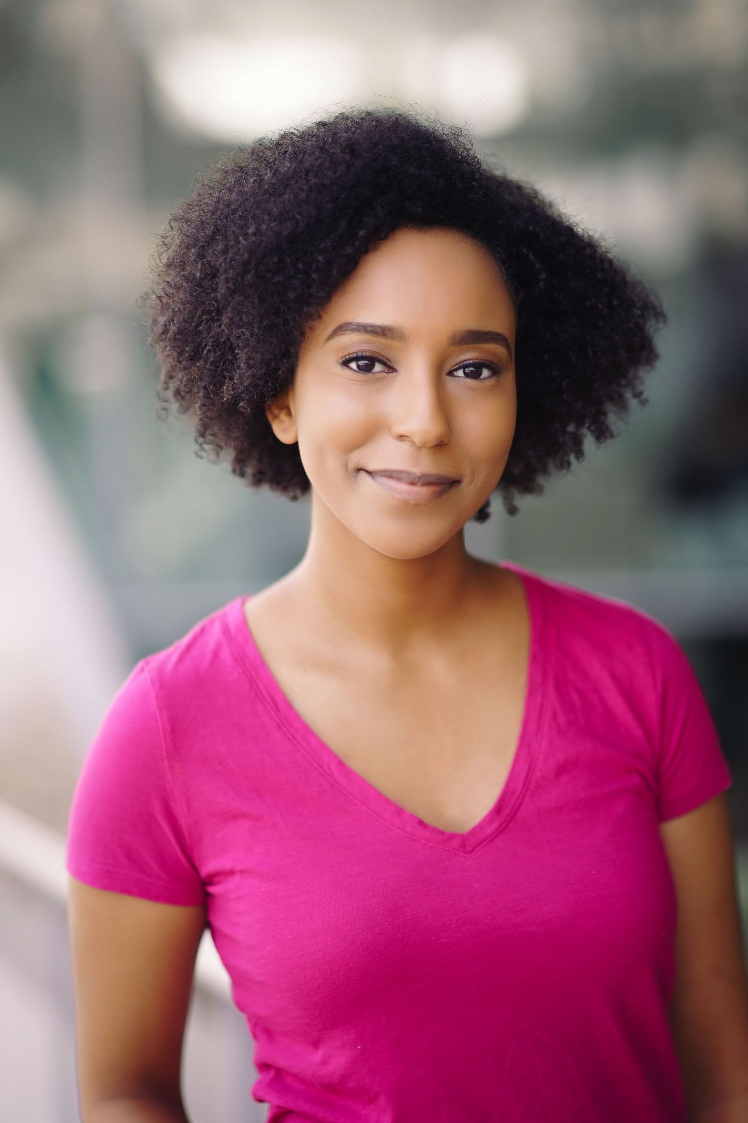 Portrait of person with natural curly hair wearing bright pink v-neck t-shirt against blurred background.