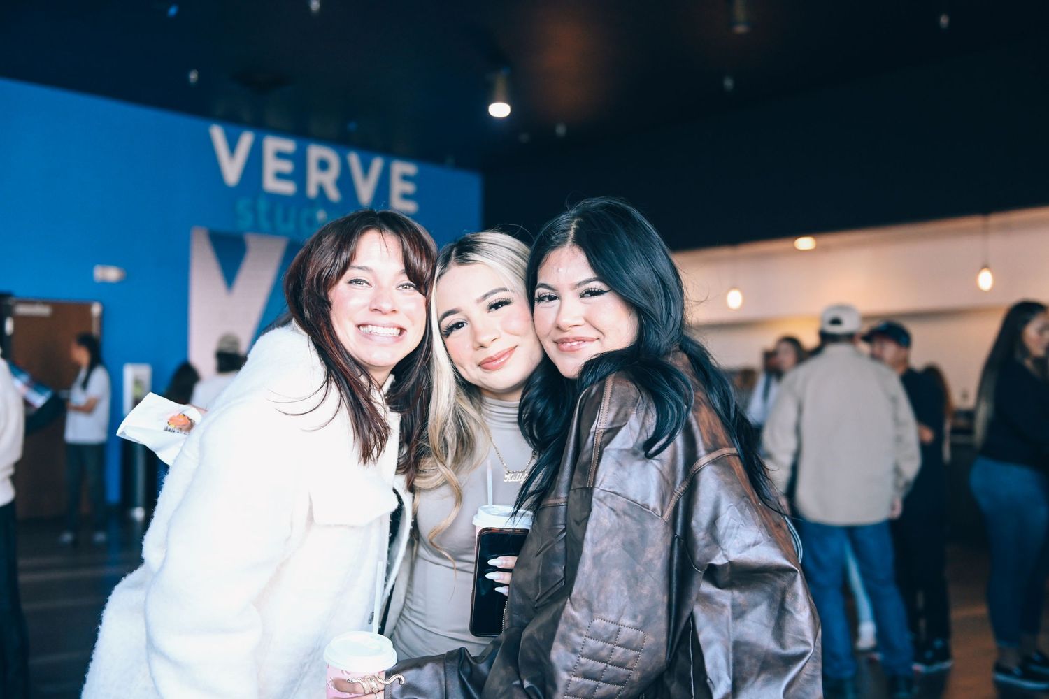 Three friends pose together at a Verve event with signage visible in the background.
