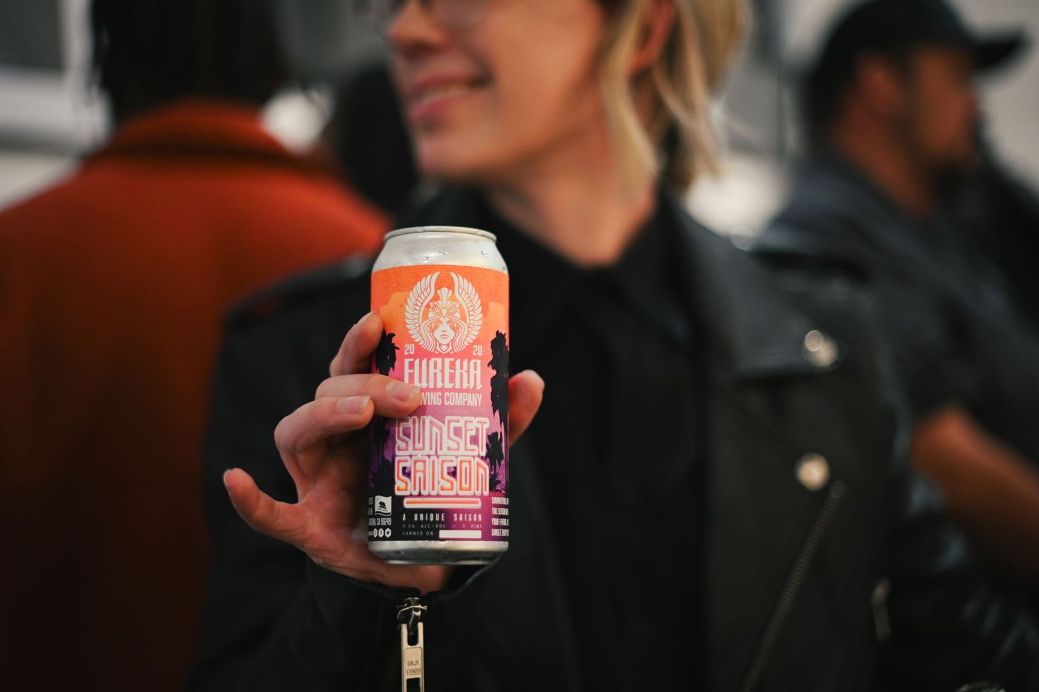 Hand holding an orange craft beer can against a blurred background at a social gathering.