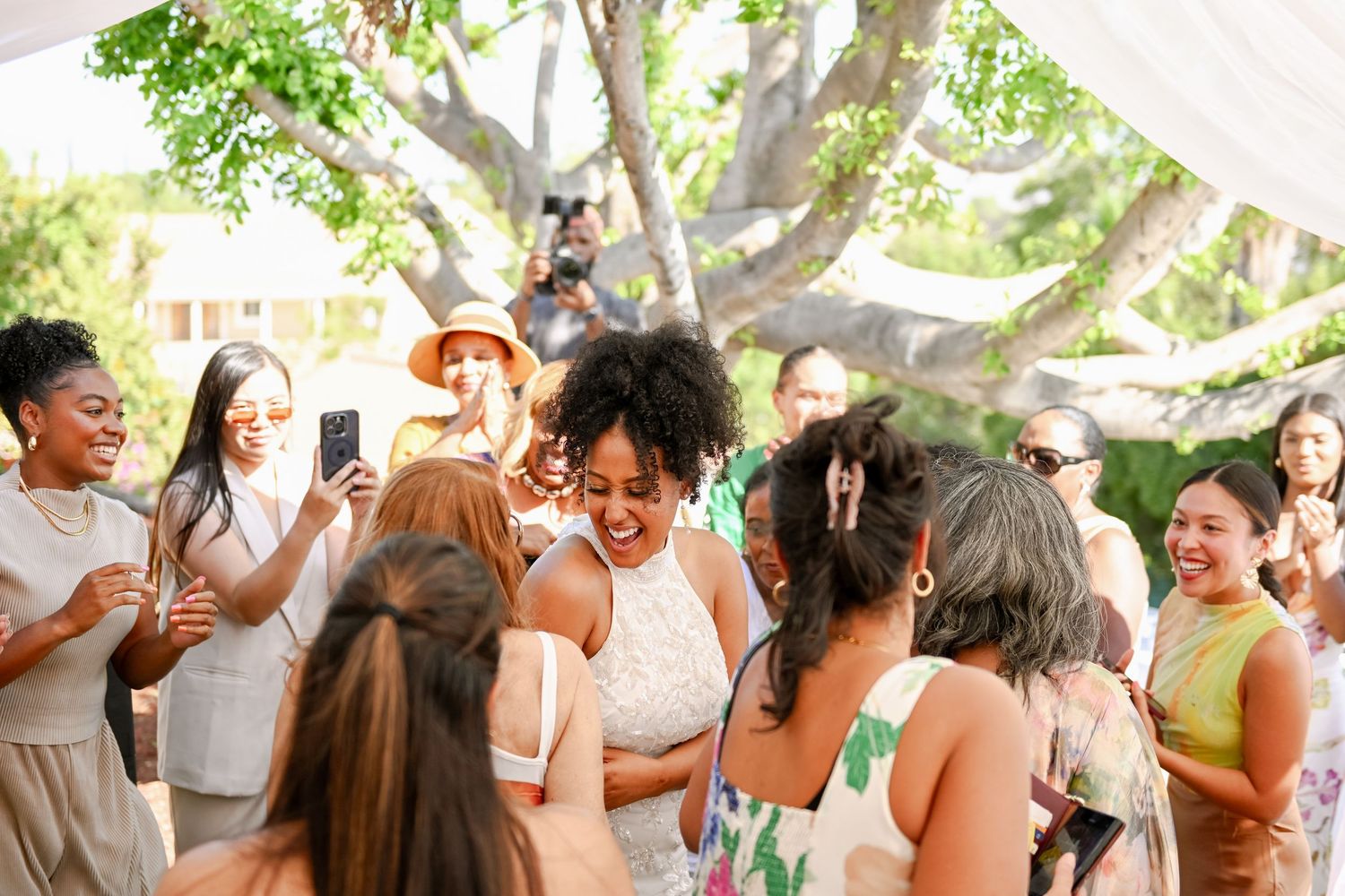 Joyful wedding celebration outdoors under trees as guests smile and take photos with smartphones.