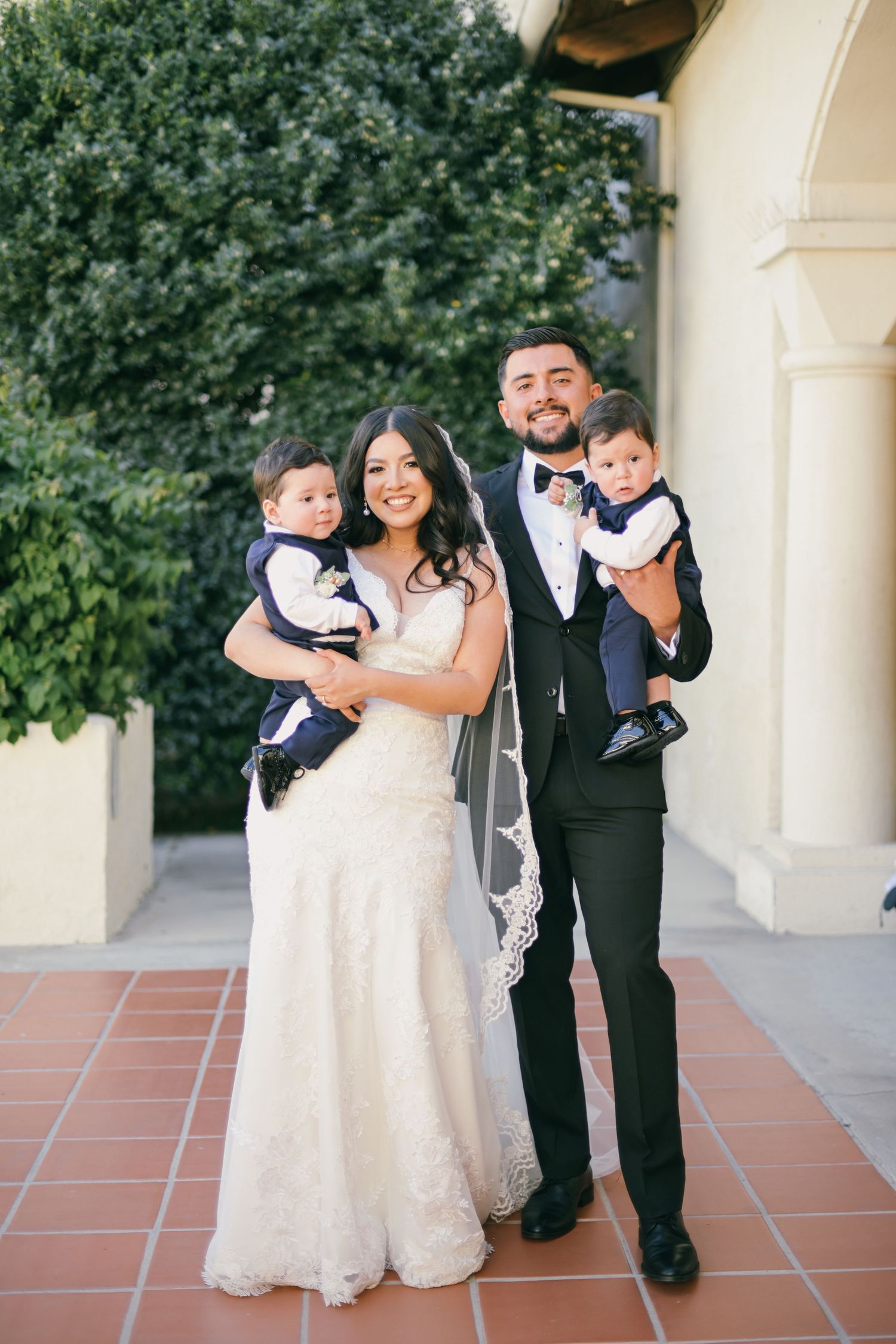 Wedding couple in formal attire holding twin babies outside church.