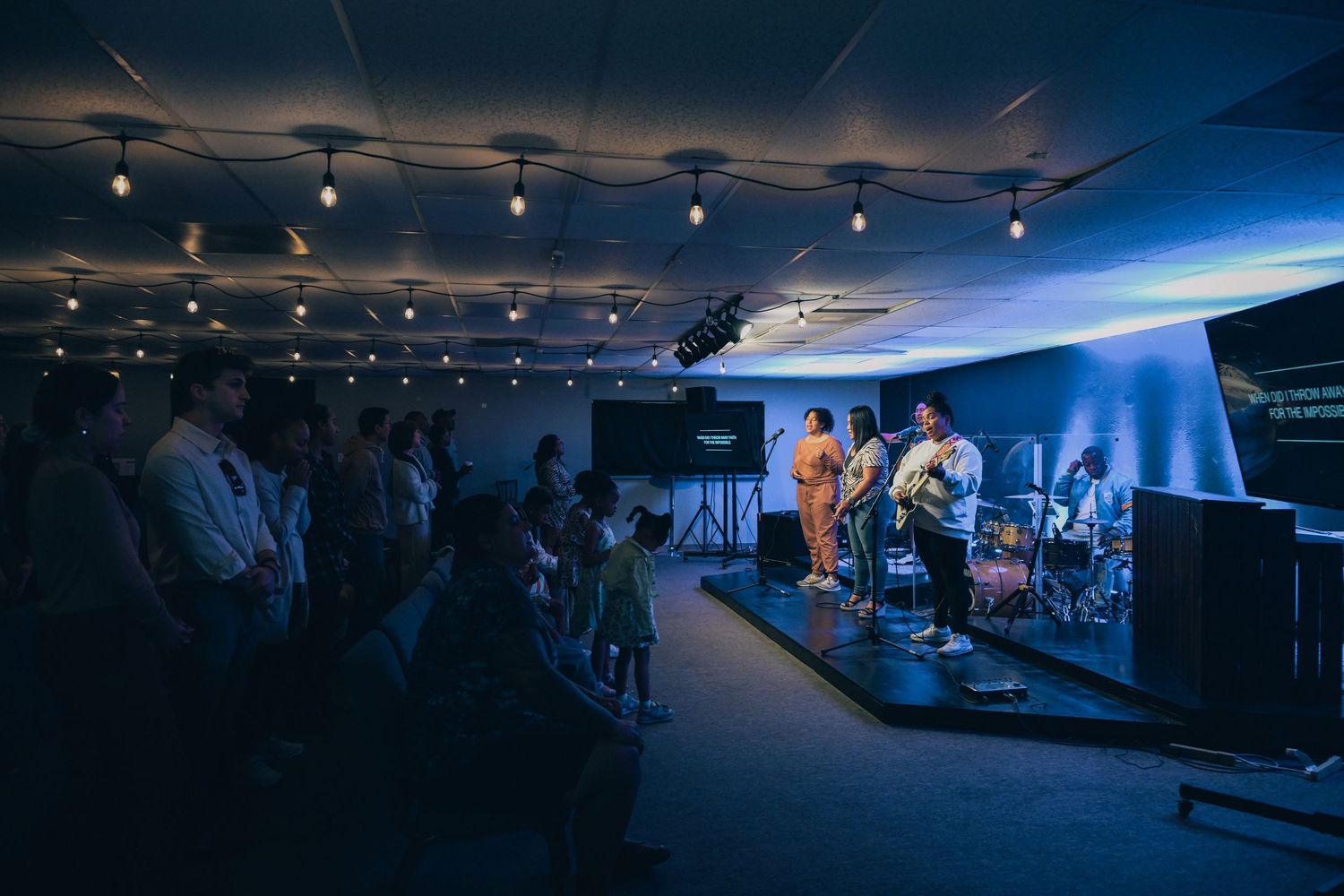 Band performs on an illuminated stage in a dimly lit venue with audience members watching from their seats.