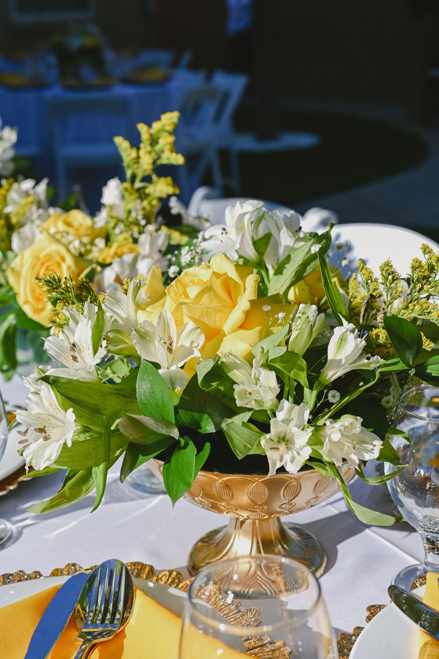 Yellow and white floral centerpiece arrangement in gold vase on white tablecloth.
