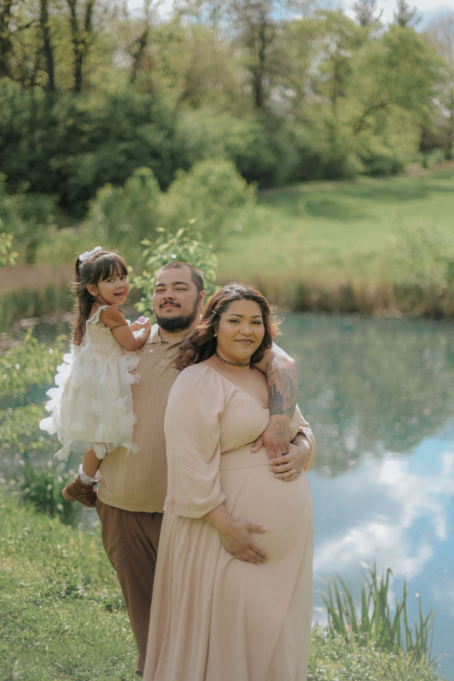 Family of three poses by a pond with trees and greenery in the background.