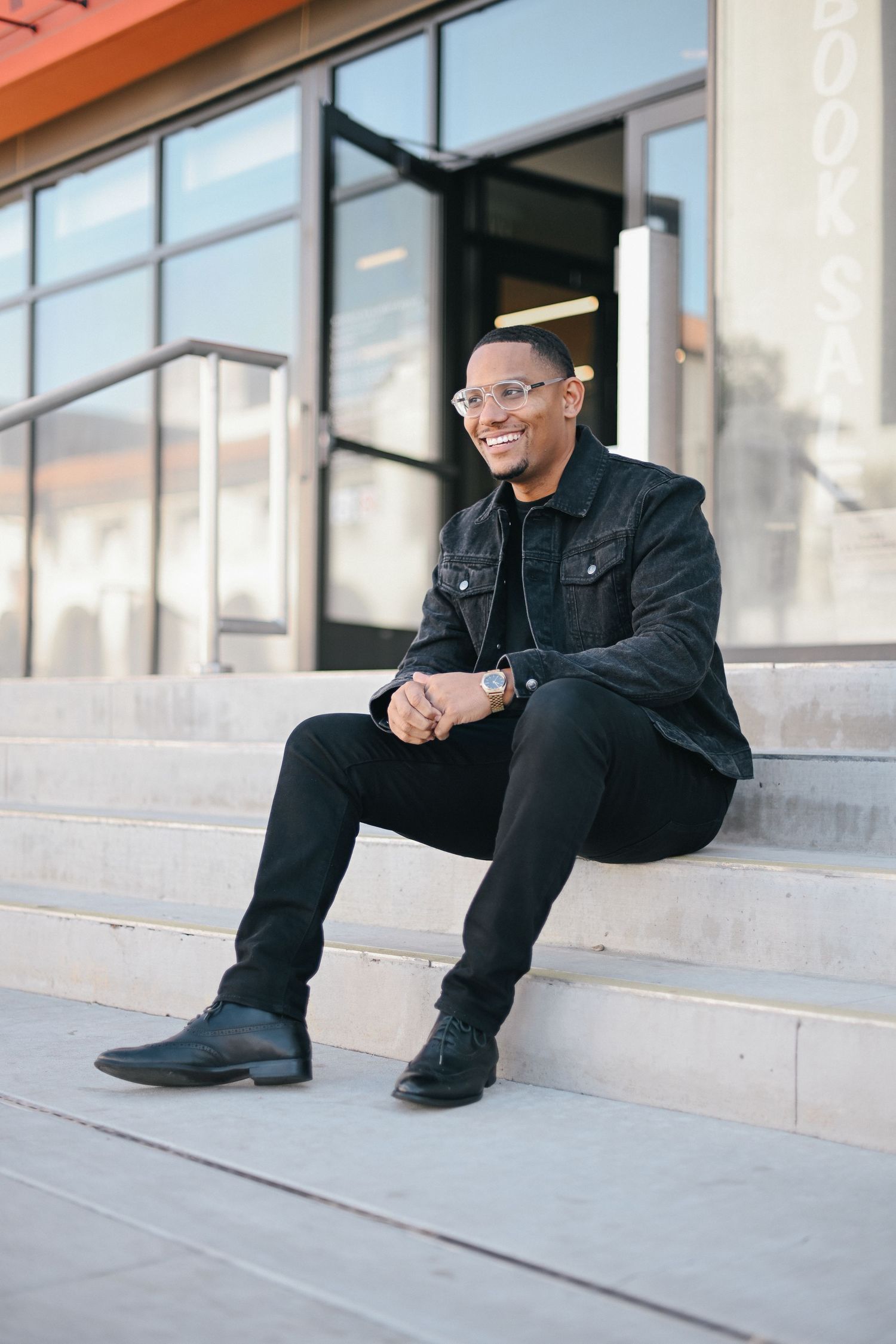 Person wearing black outfit and glasses sitting on concrete steps outside a building.