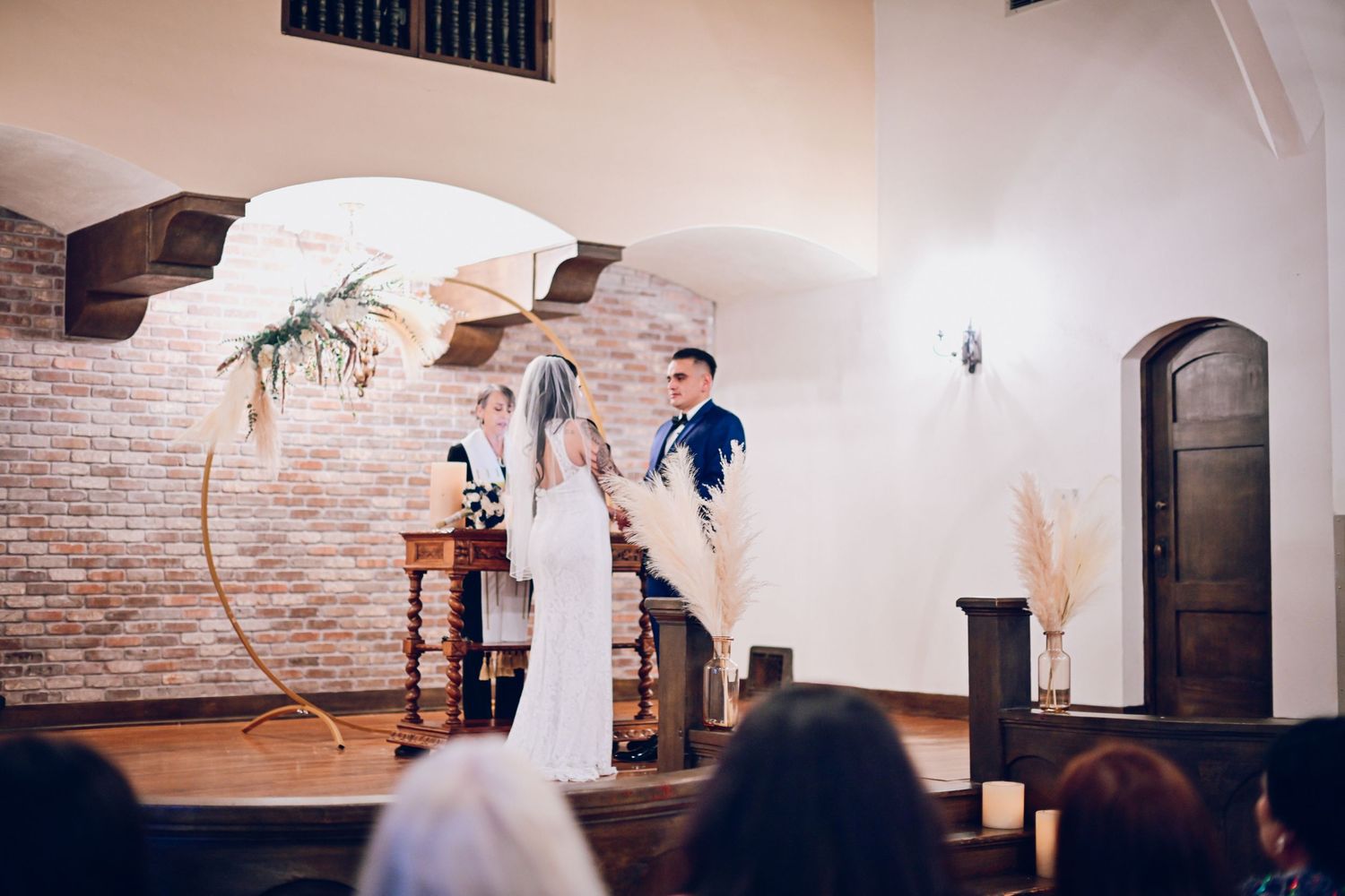 A wedding ceremony takes place under an elegant circular arch in a brick-walled venue.