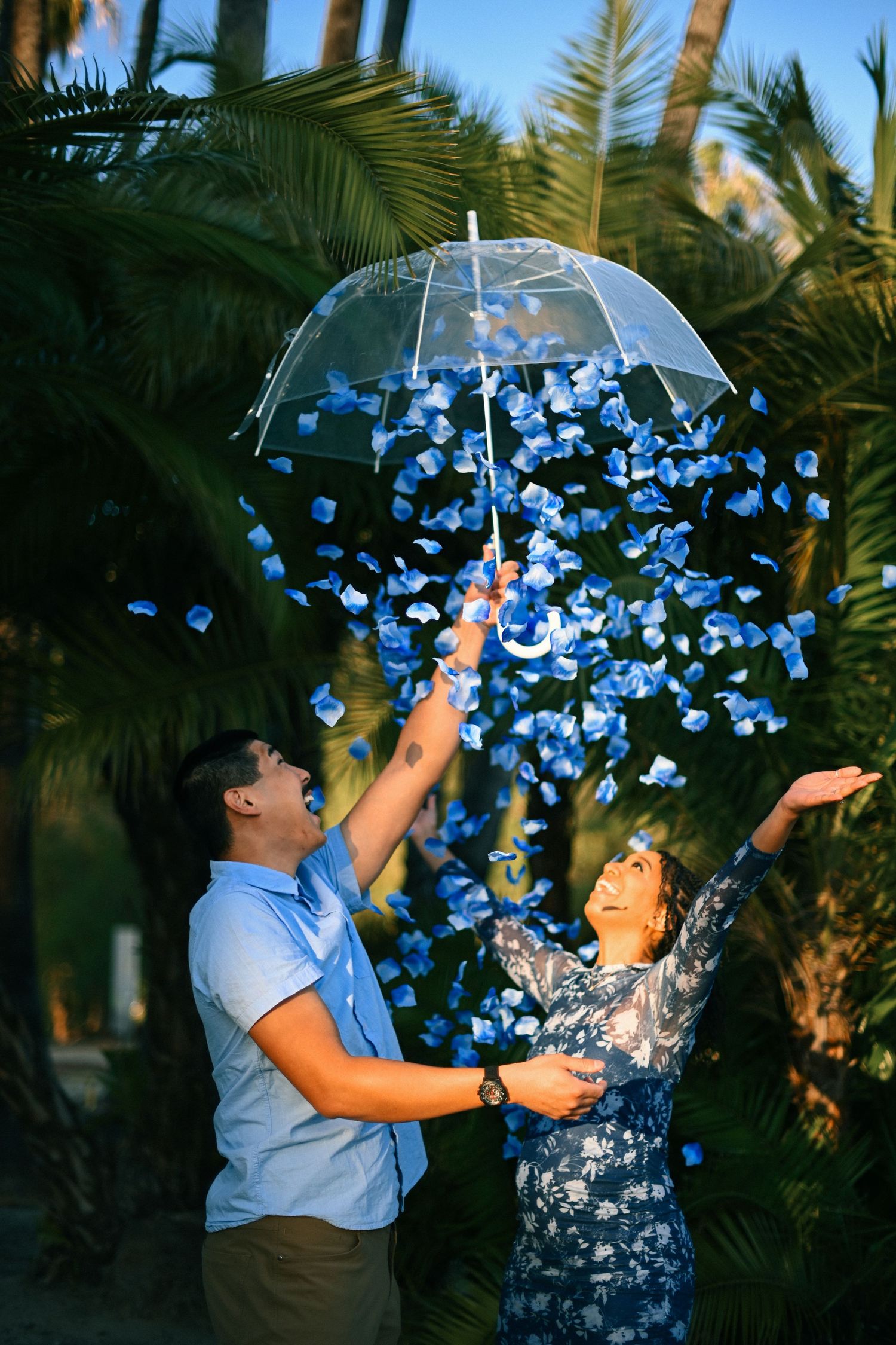 A couple celebrates with blue confetti falling from a clear umbrella among palm trees.