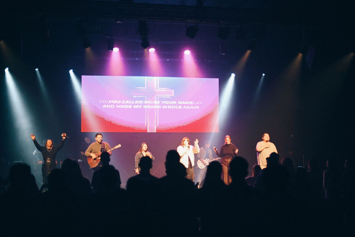 Worship band performing on stage with colorful spotlights and projected lyrics.