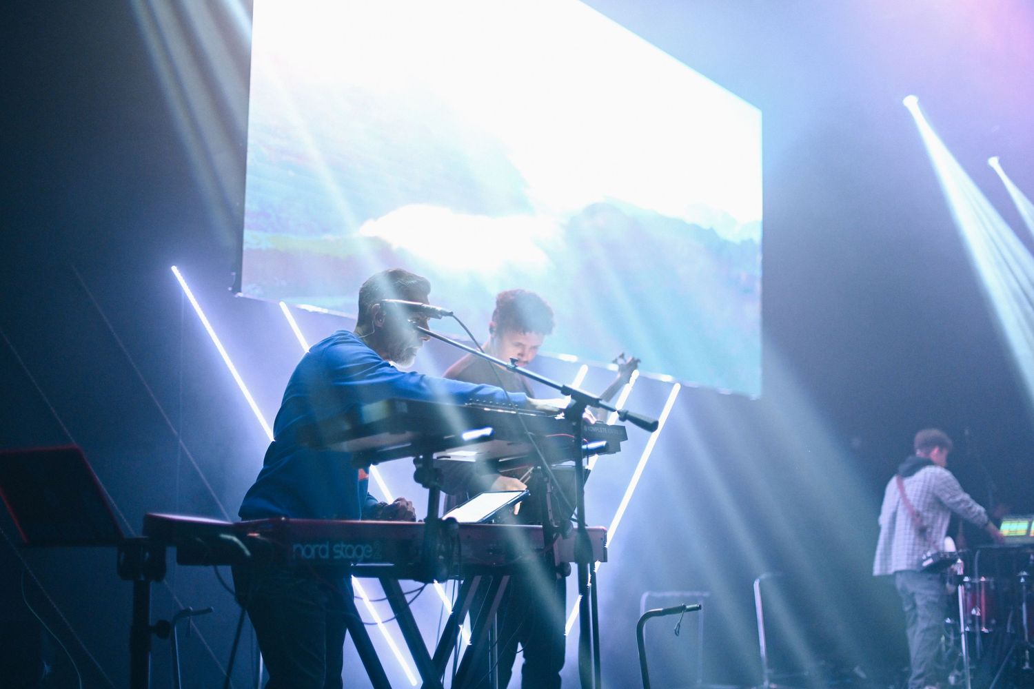 Musicians perform on stage with dramatic lighting and electronic instruments during a live concert performance.