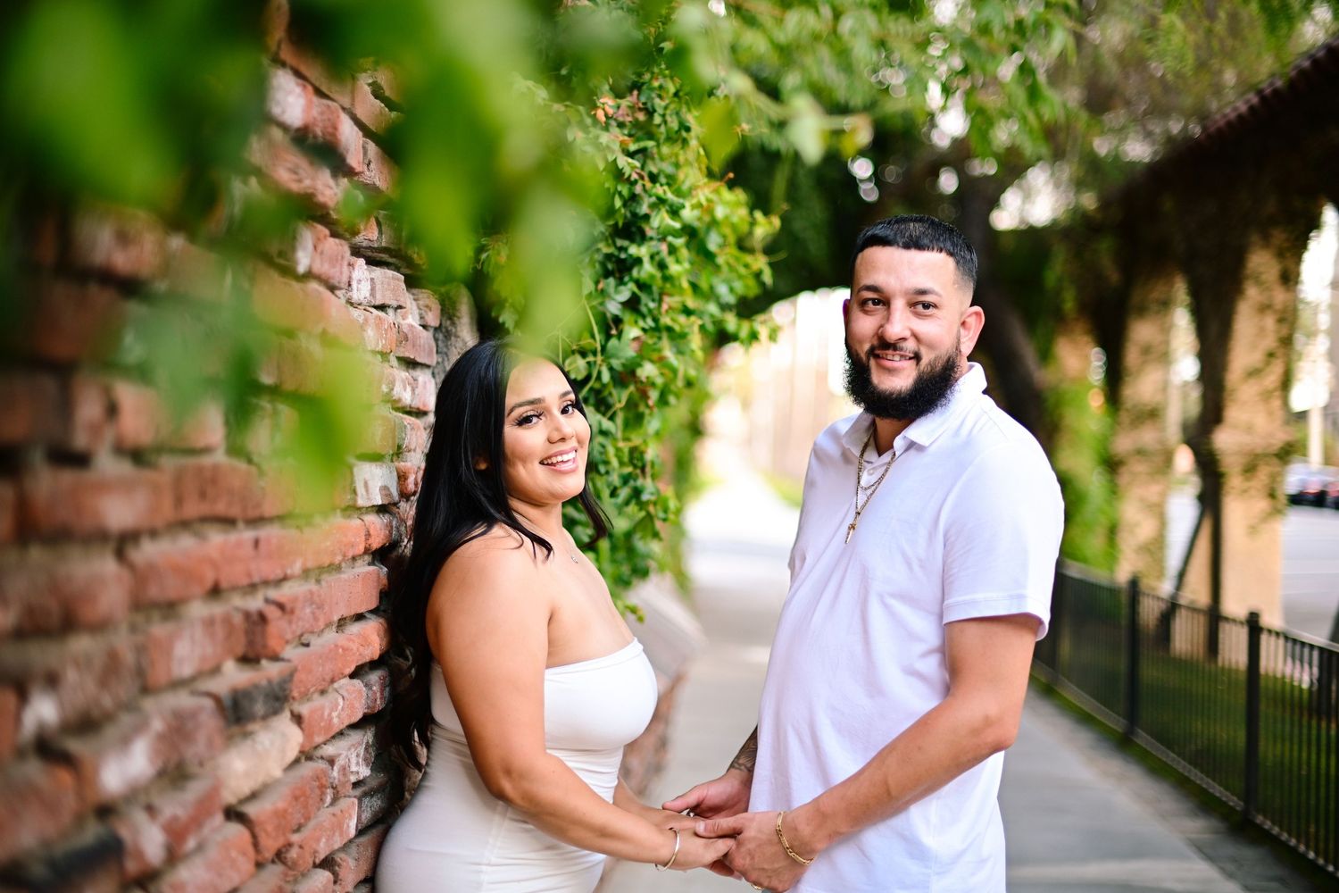 Engaged couple sharing romantic moment against brick wall with greenery.