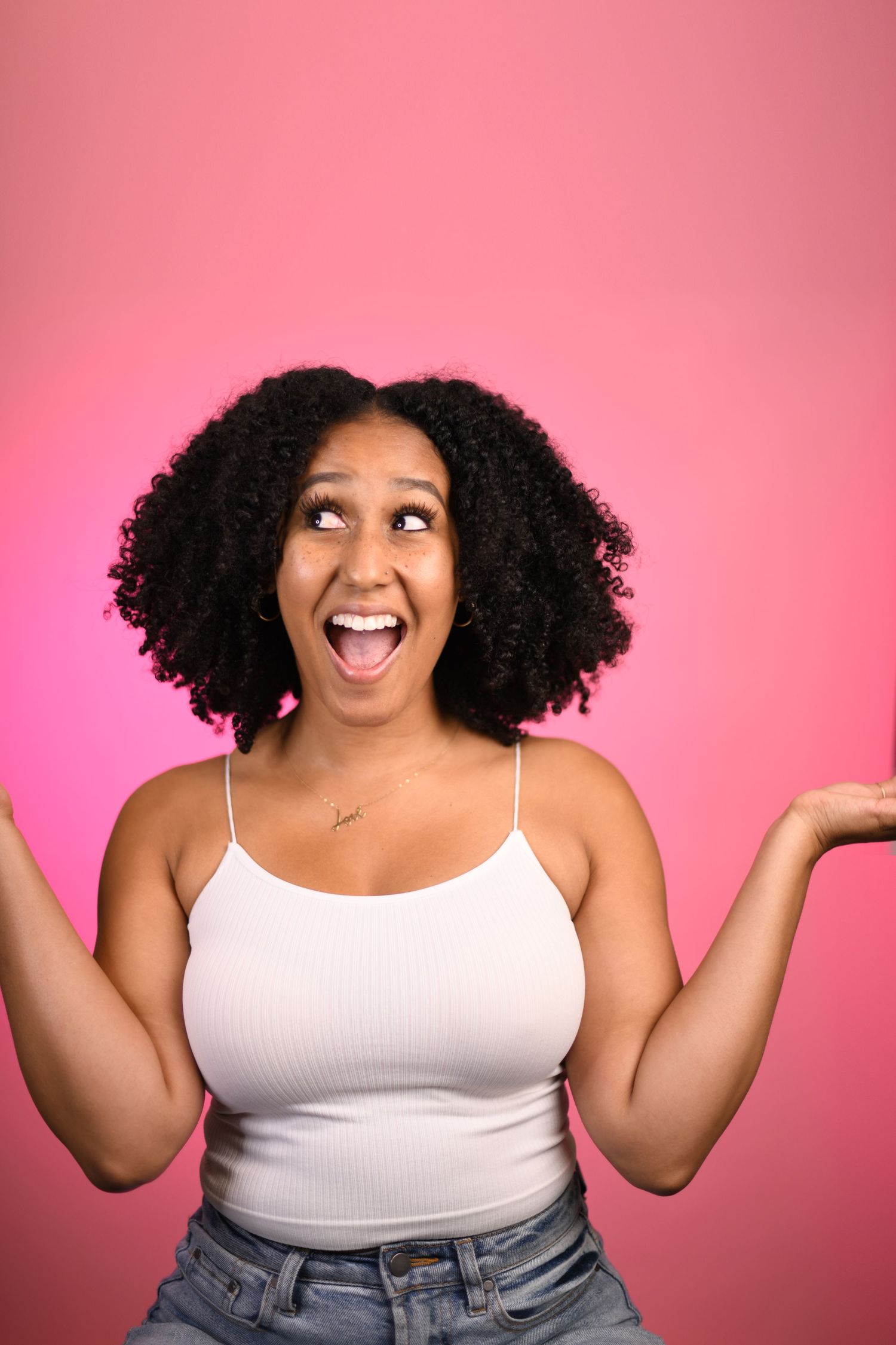 Joyful person with curly natural hair laughing expressively against pink background while wearing white tank top.