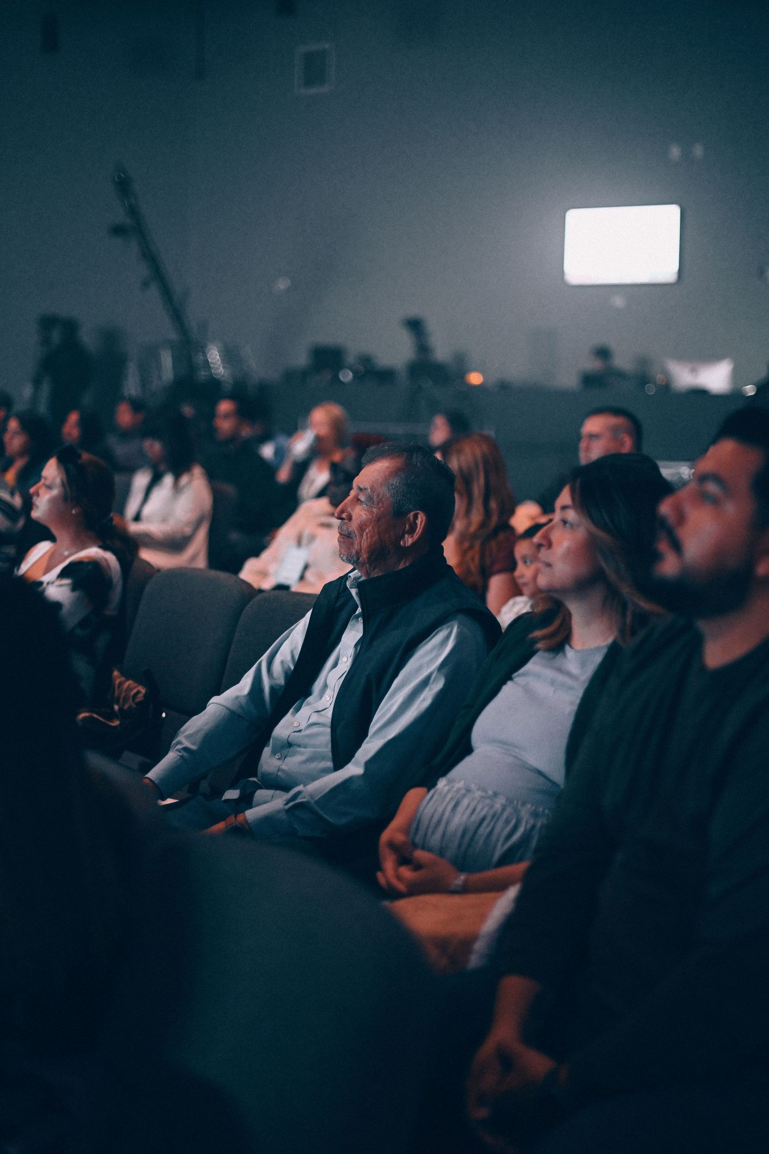 Audience members watch attentively in a dimly lit theater or auditorium.