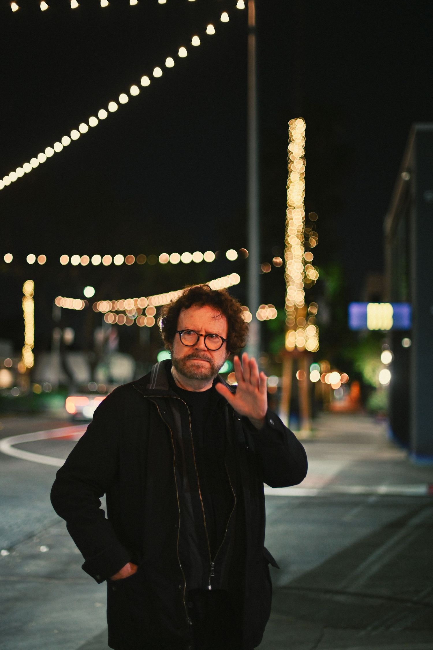 Person in winter attire waves at camera on city street with string lights in background.
