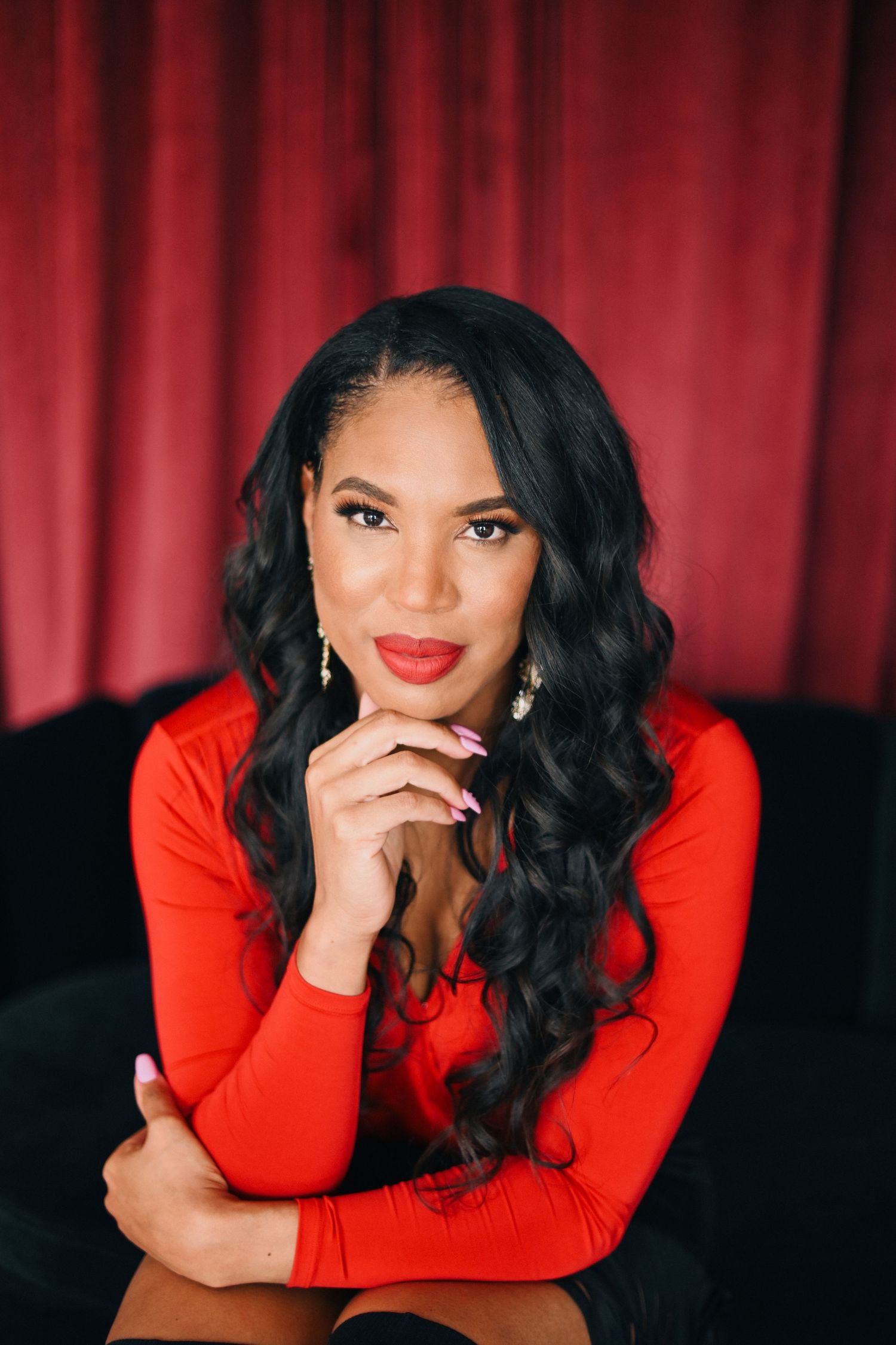 Portrait of a person in a red sweater posing with hand under chin against red curtains.