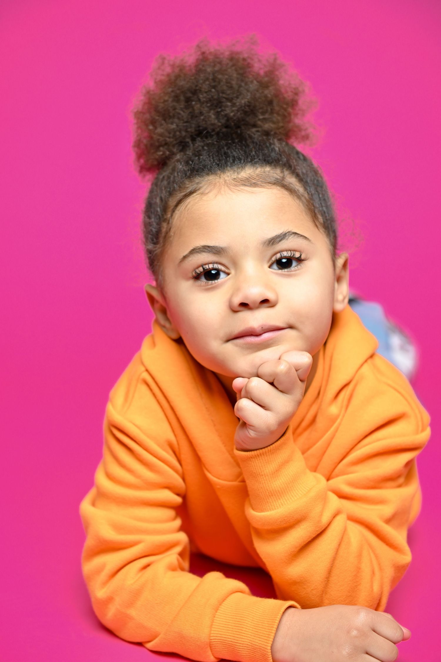 Portrait of child in orange hoodie against pink background with natural hair in bun style.