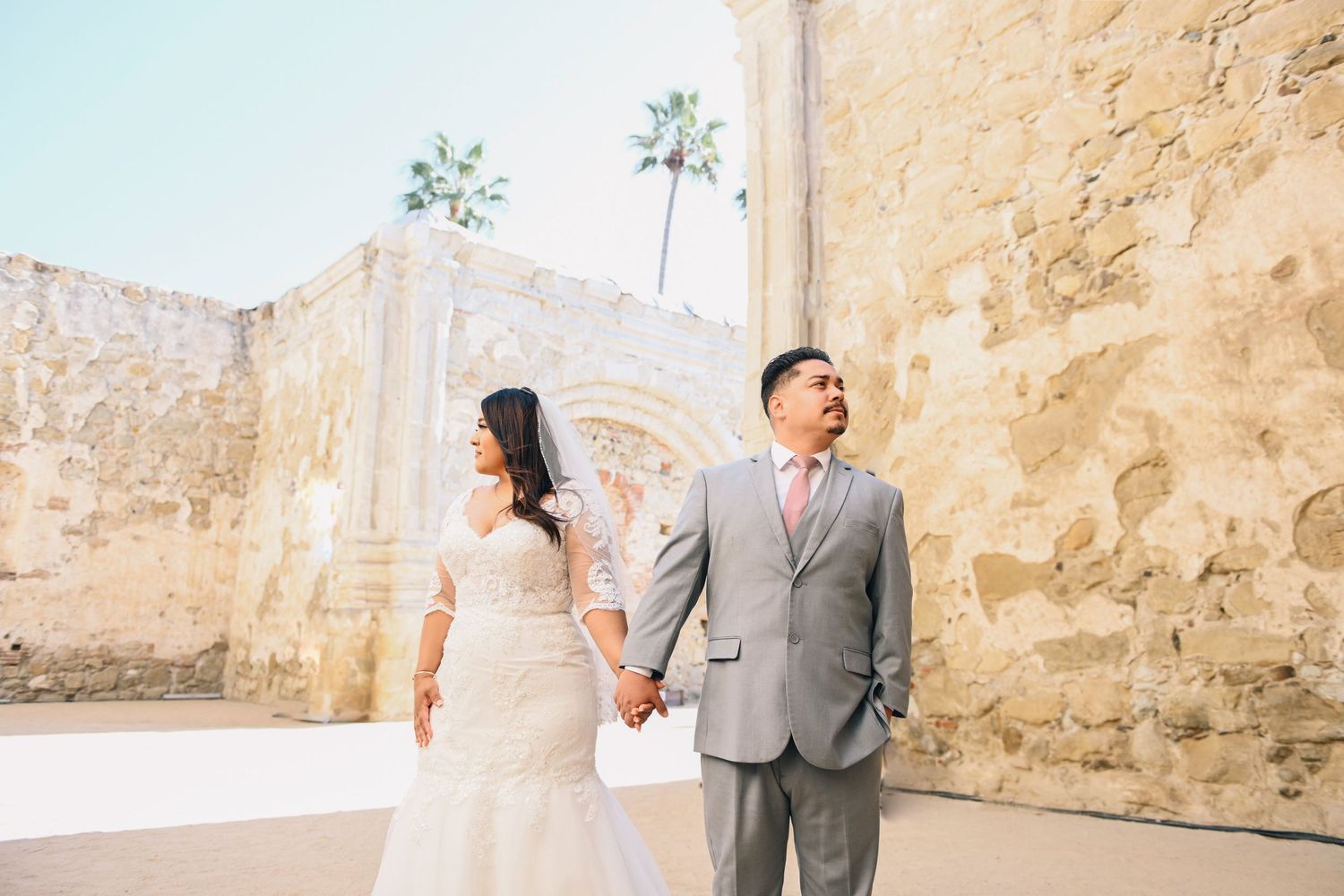 Newlyweds hold hands near stone wall with palm trees in background.