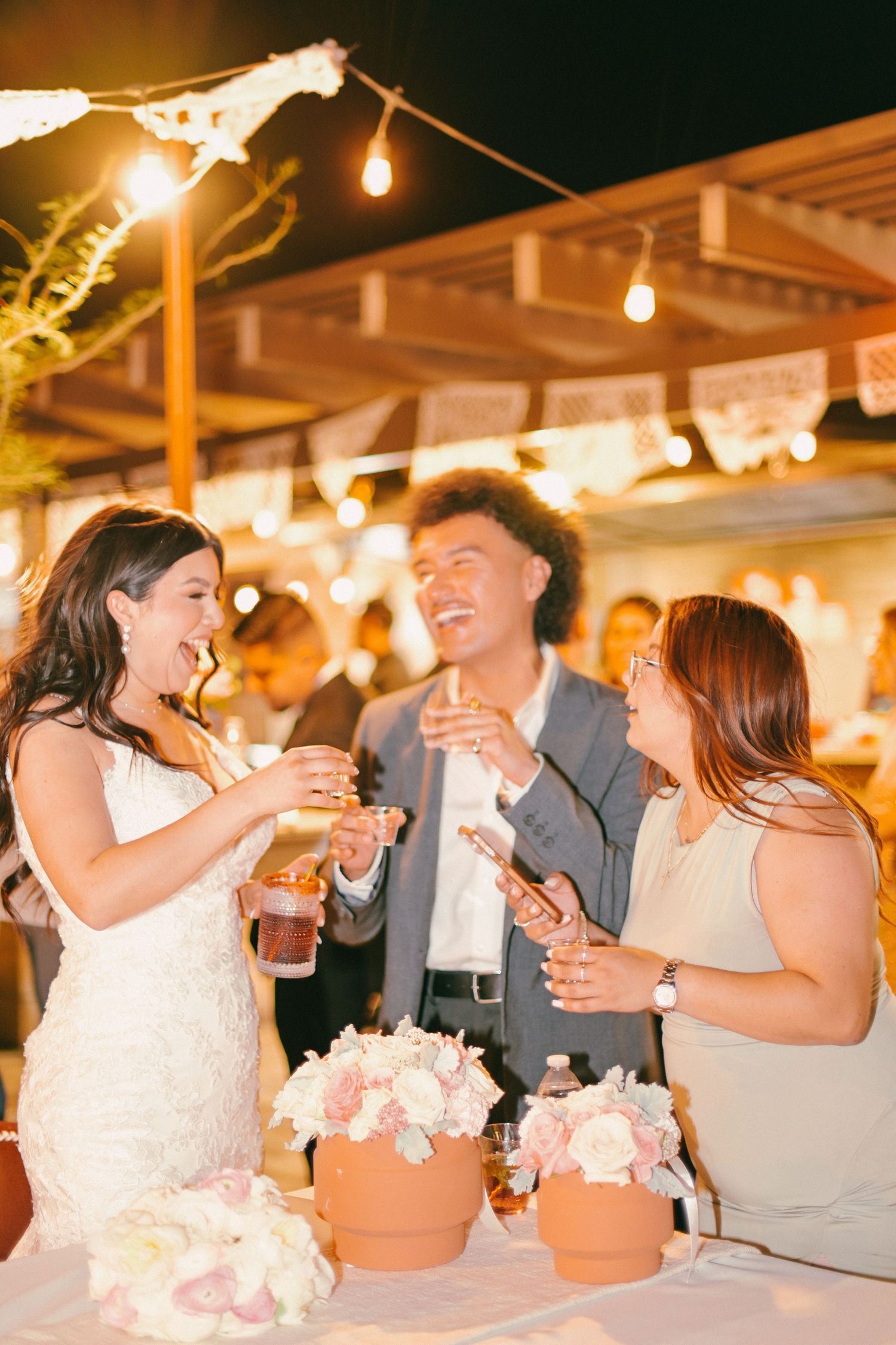 Wedding guests laugh and celebrate at outdoor reception under string lights.