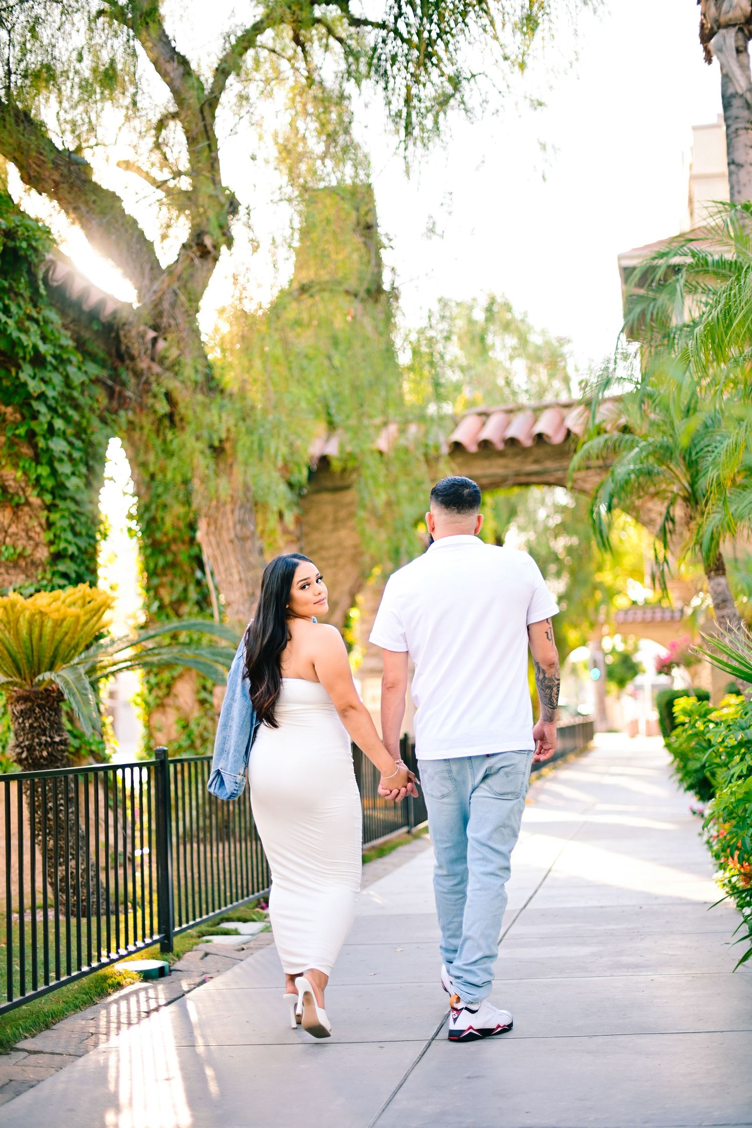 Couple walking hand in hand along scenic path with Spanish architecture and hanging trees.