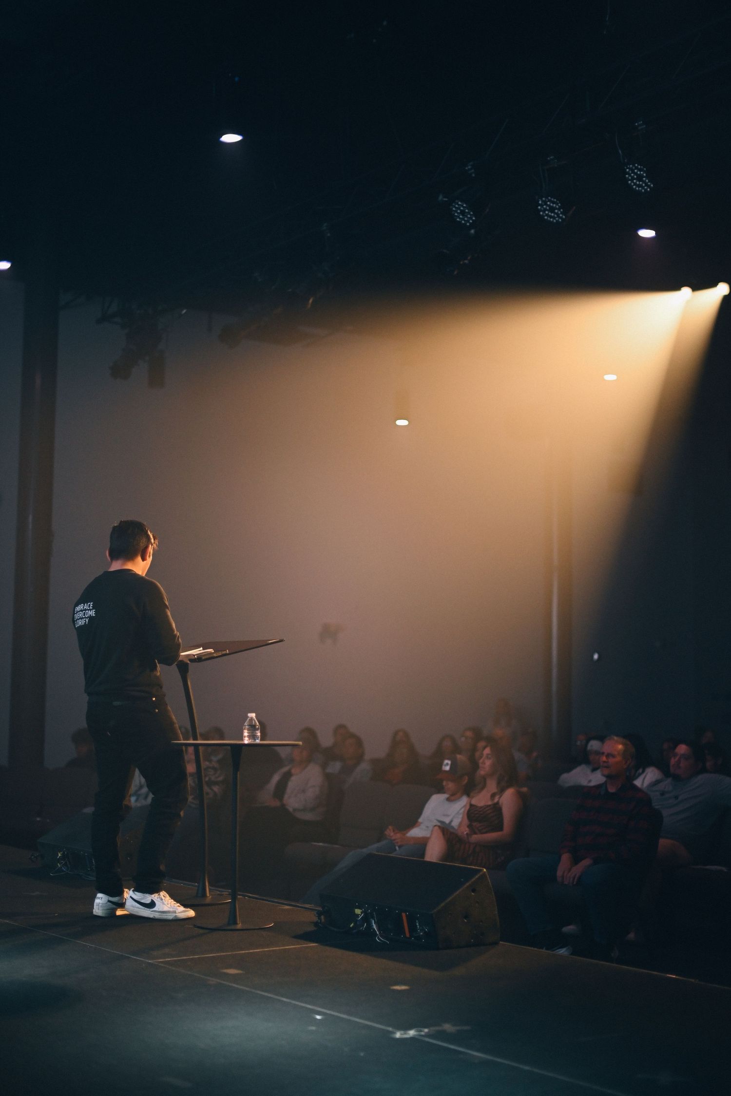 A speaker on stage addresses an audience under dramatic spotlight in a dark theater setting.