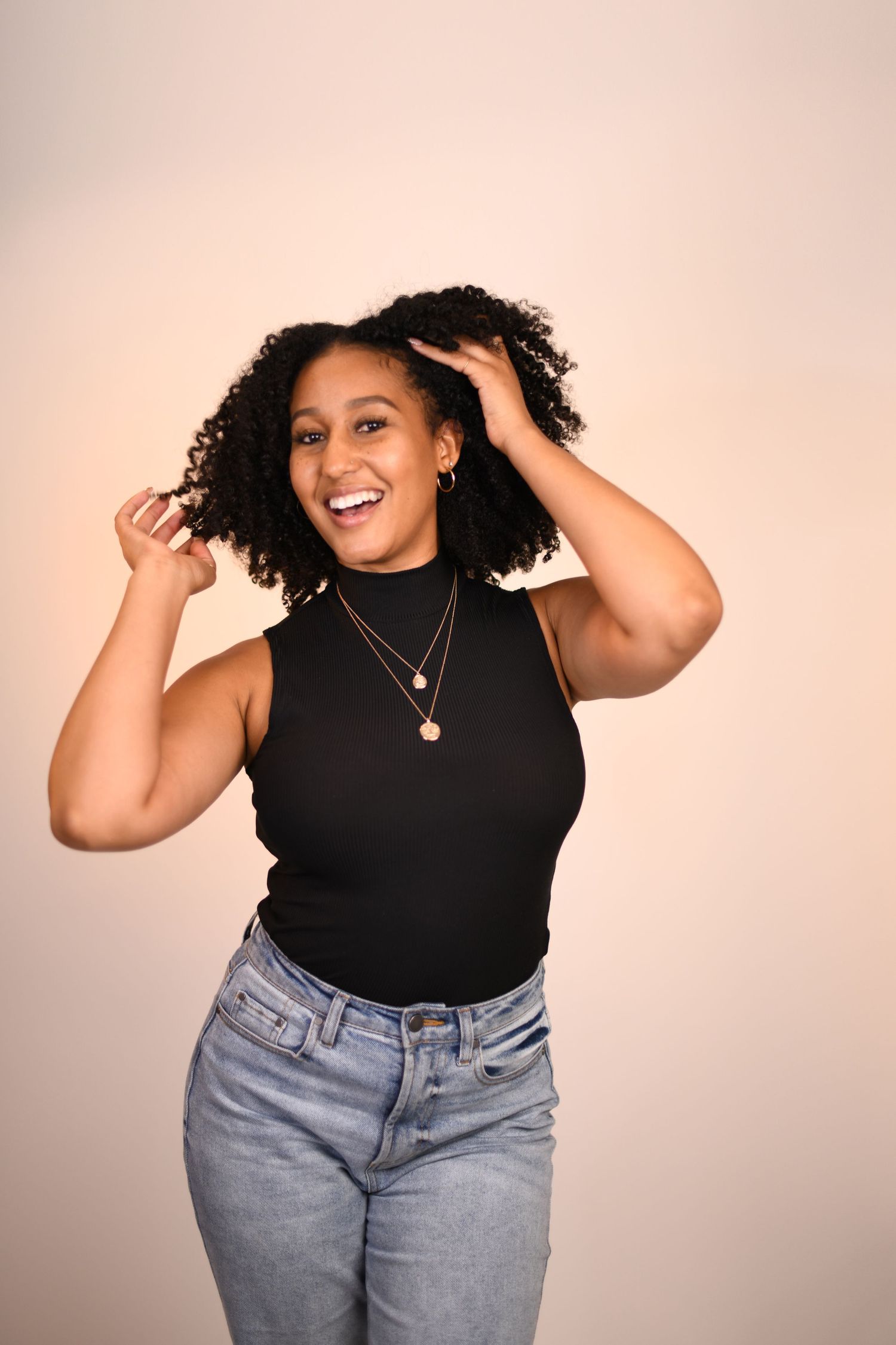 Joyful person with curly hair wearing black top and jeans against pink background.