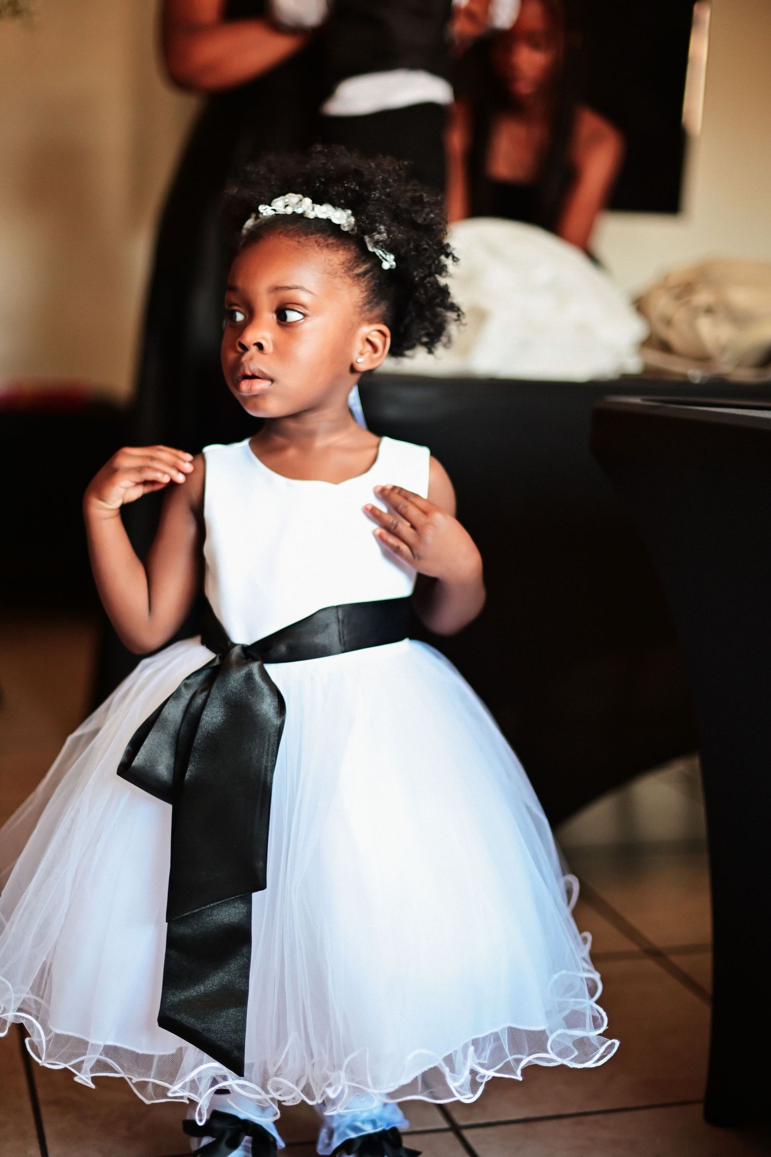A young flower girl in a white tulle dress with black sash poses at a wedding.