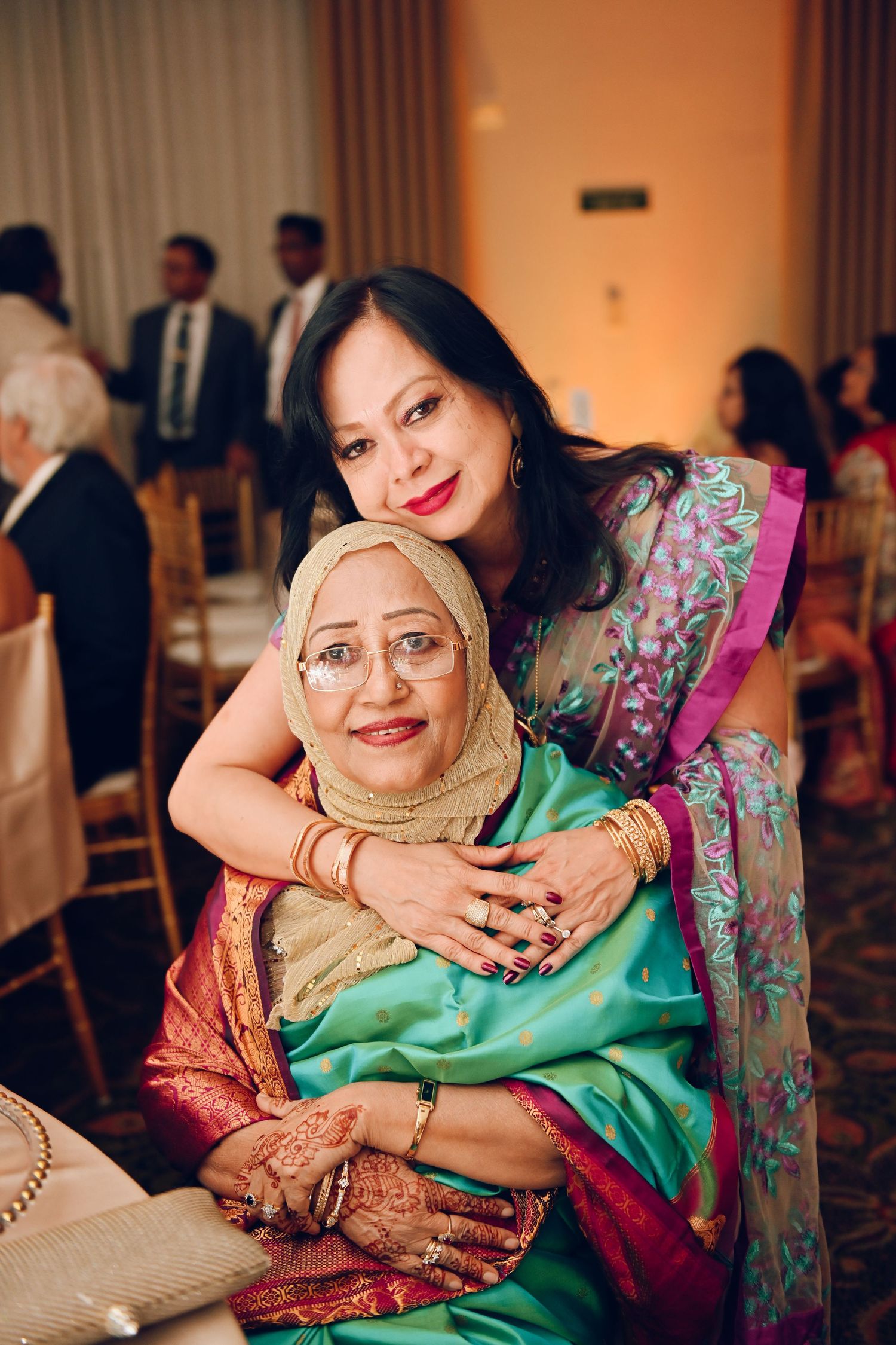 Two women in colorful traditional South Asian attire sharing an embrace at a formal event.