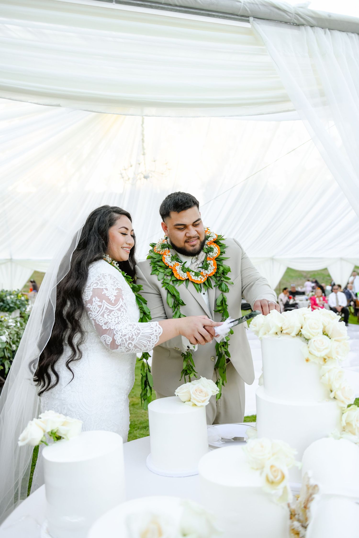 Couple cutting their wedding cake together under a white tent with traditional Hawaiian leis.