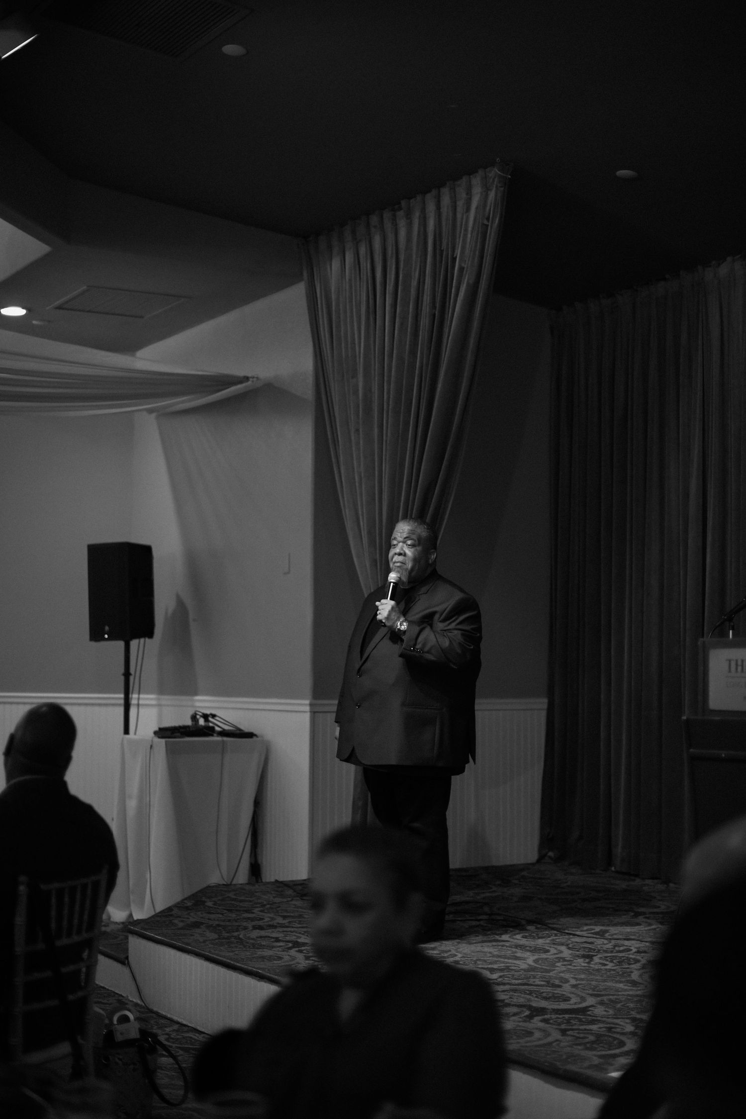 A speaker stands at a podium during an indoor event with curtains and dim lighting in the background.