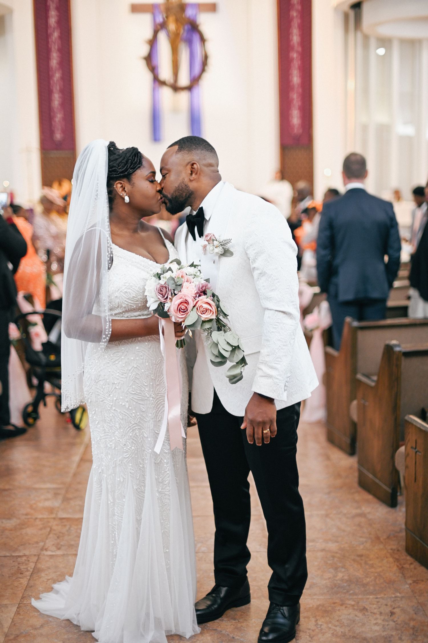 Newlyweds share romantic kiss in church aisle with bride holding pink floral bouquet.