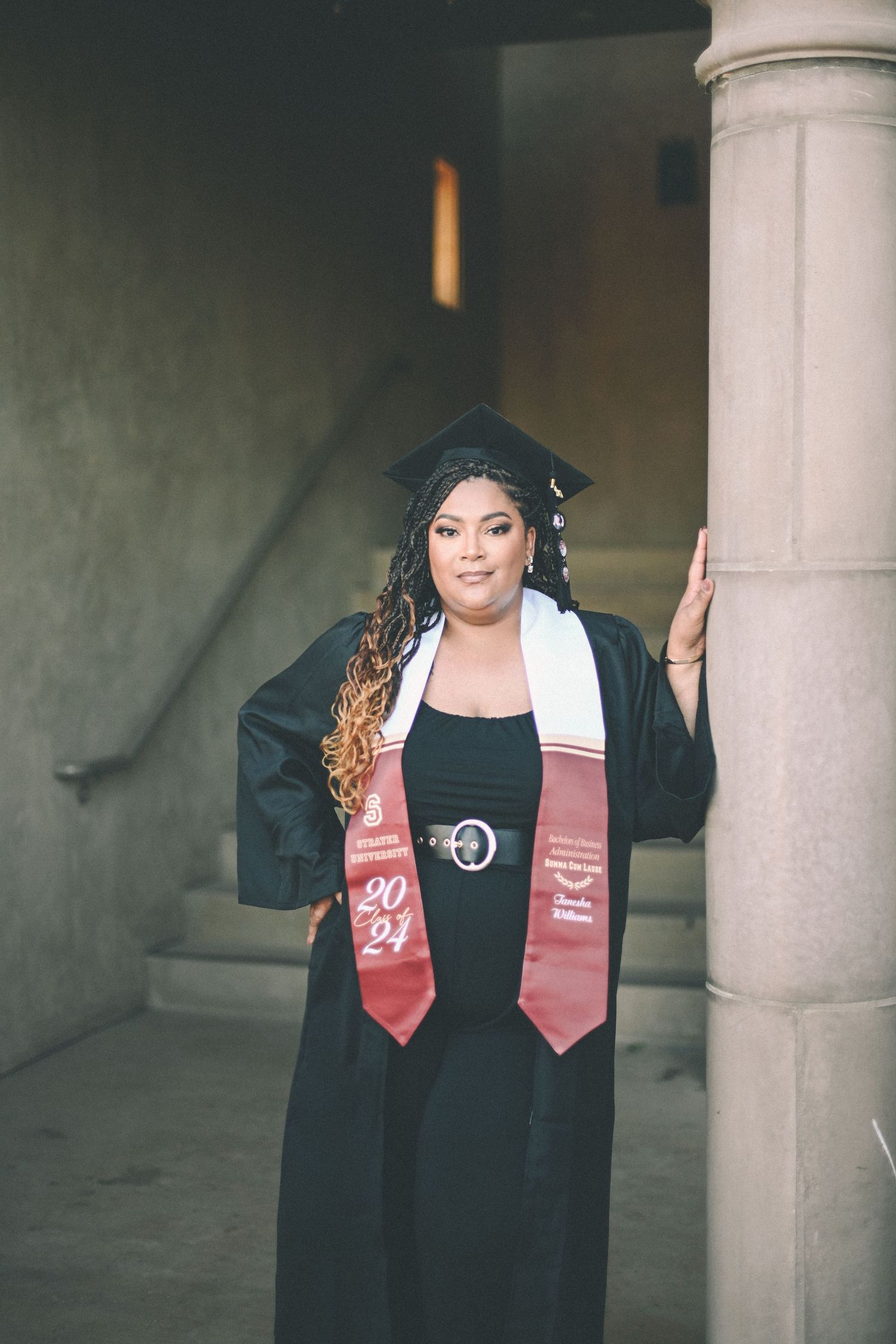 Graduate in black cap and gown with red stole poses confidently by concrete pillar.