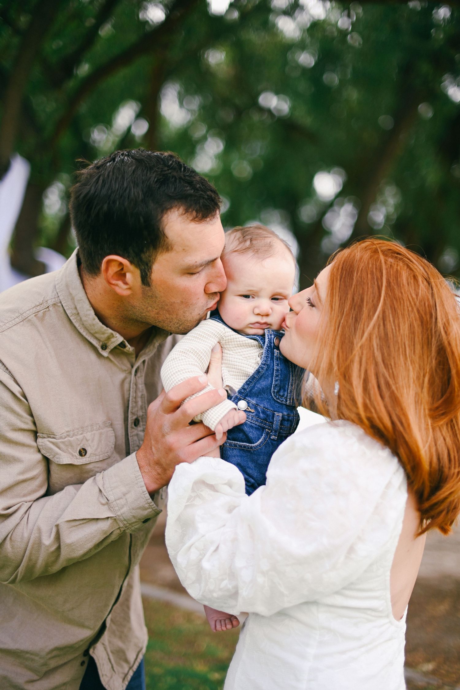Parents sharing tender moment kissing their baby in denim overalls outdoors.