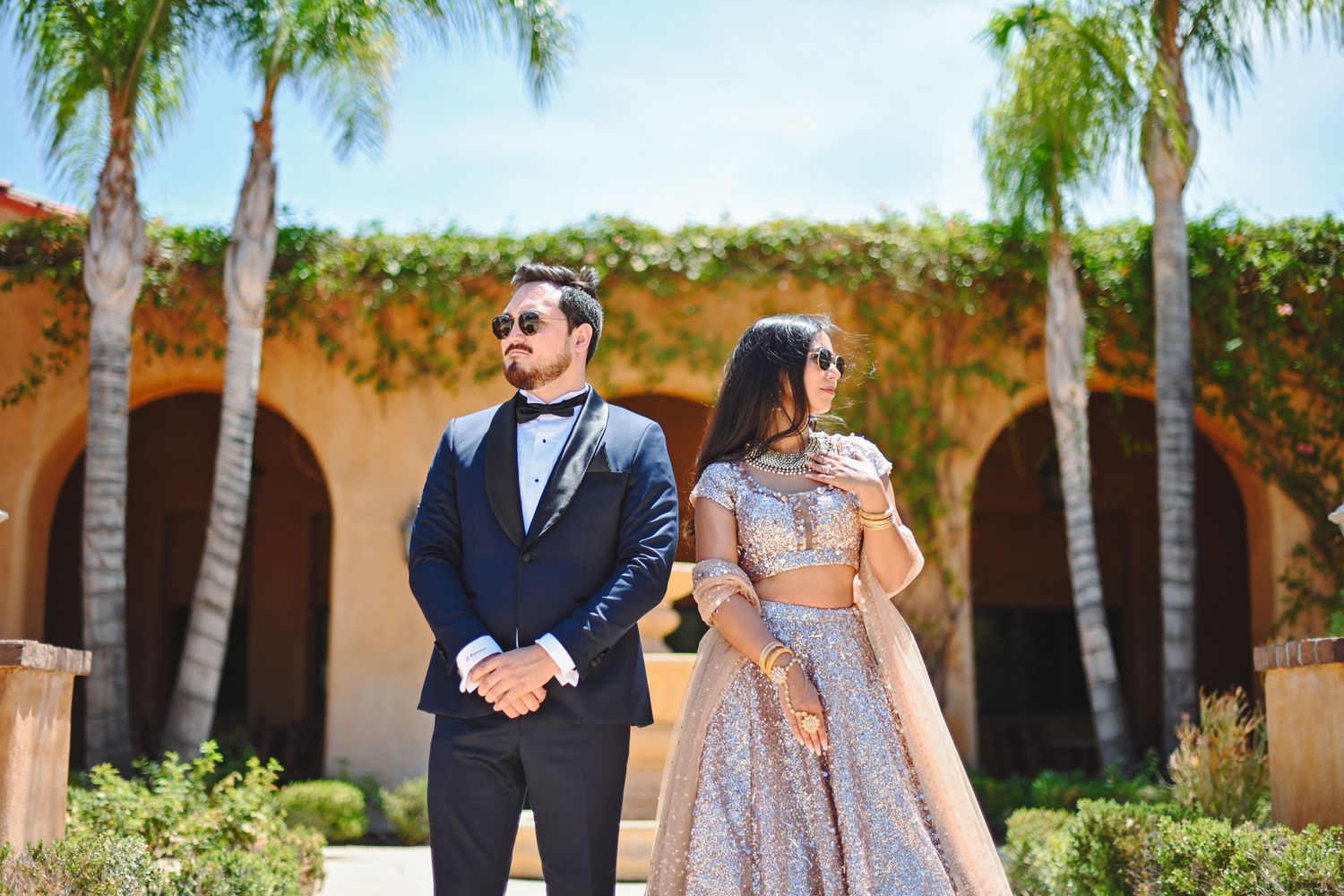 Wedding couple standing back to back in elegant attire against palm tree courtyard.