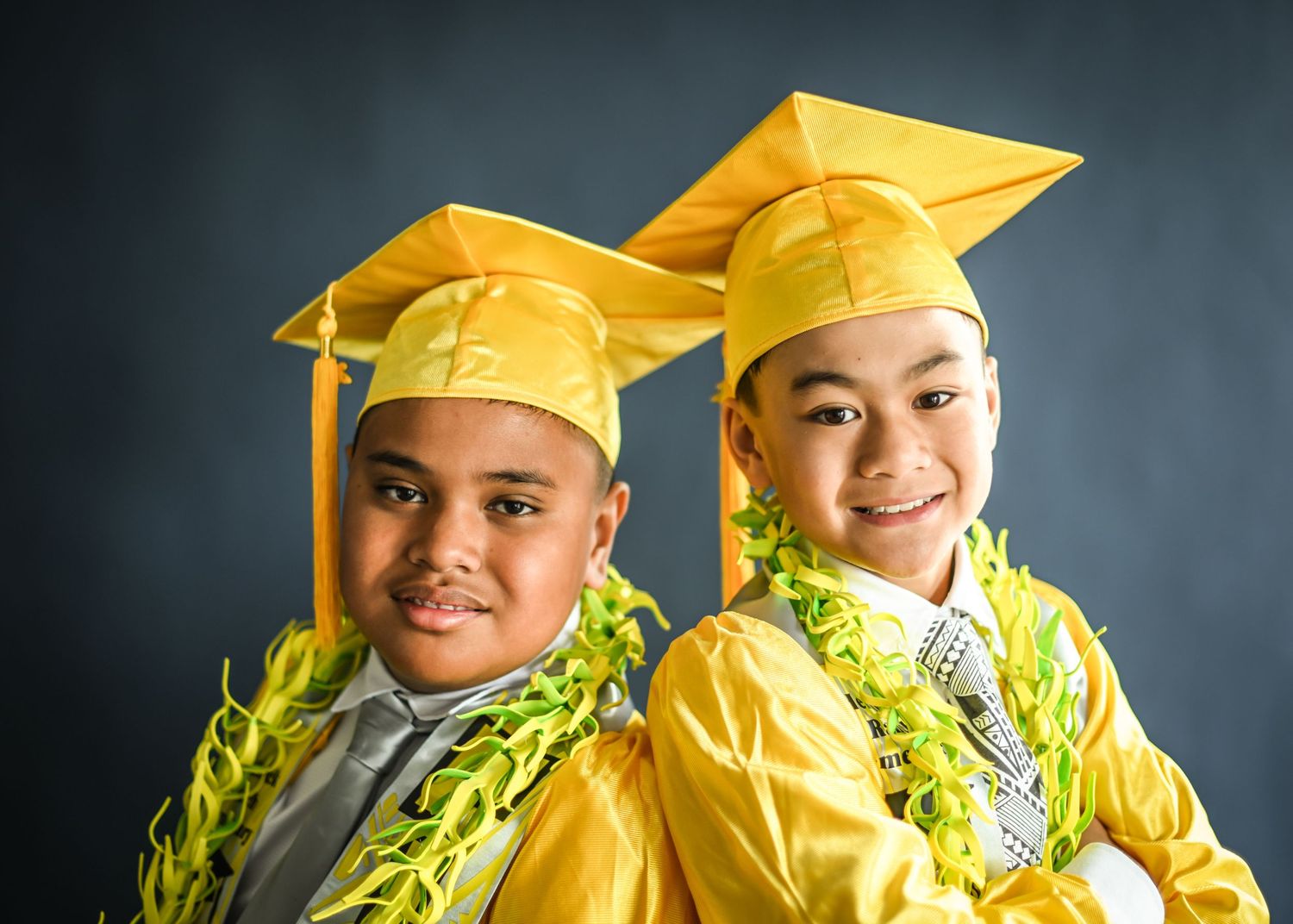 Two graduates wearing bright yellow caps and gowns with green leis smile together against a dark background.