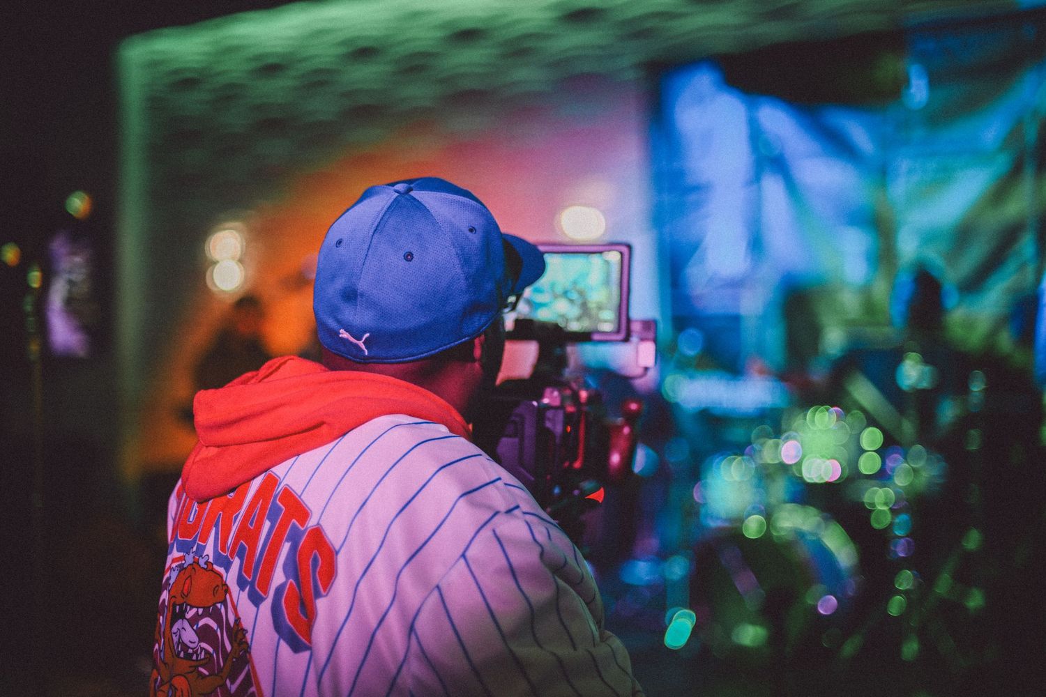 Videographer wearing baseball uniform filming with professional camera equipment in colorful lighting.