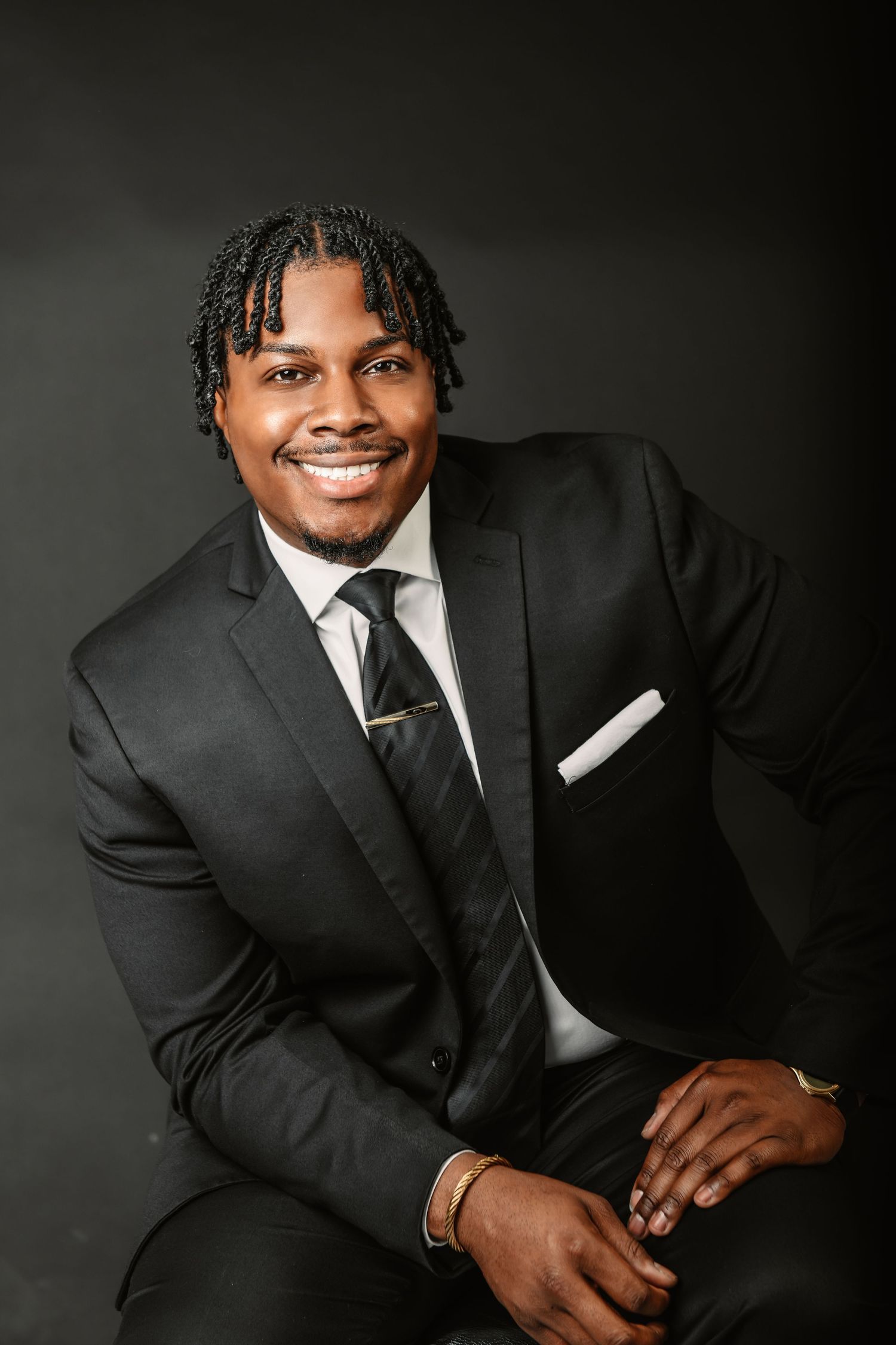 Professional headshot of person in black suit and tie smiling warmly against dark background.