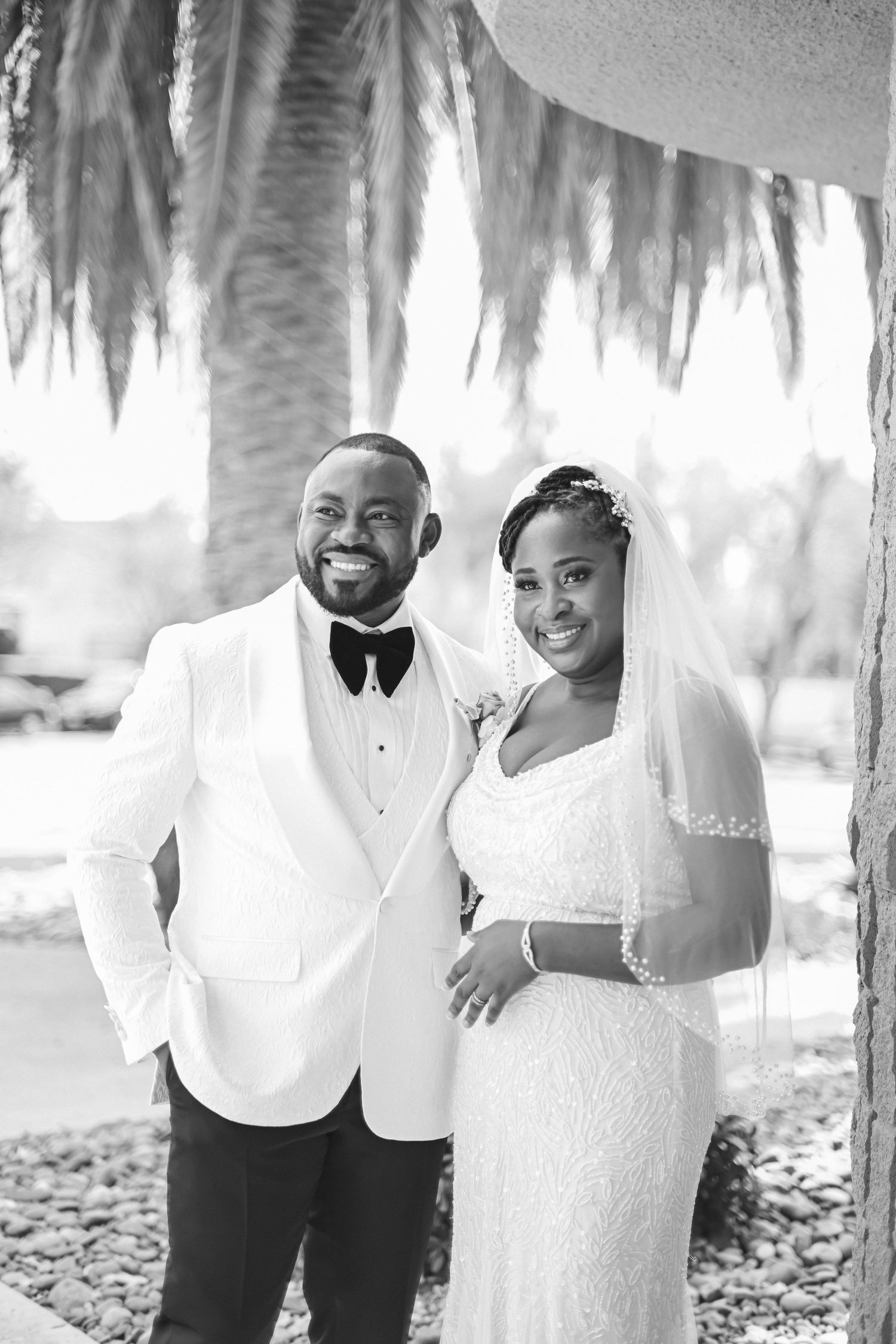 Black and white wedding photo of newlyweds in elegant attire posing under decorative hanging foliage.