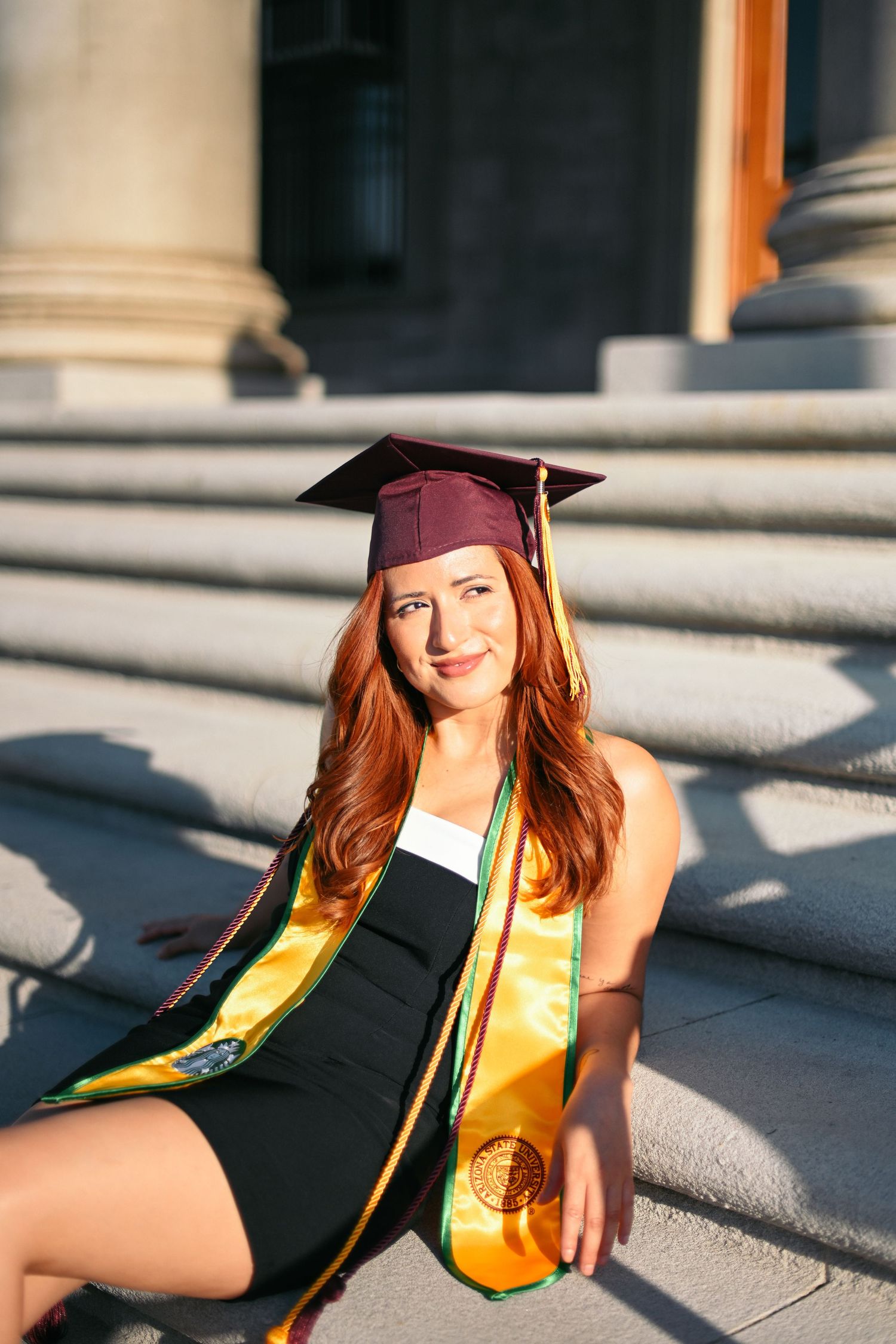 Graduate in maroon cap and gold stole sits on concrete steps during sunset graduation photoshoot.