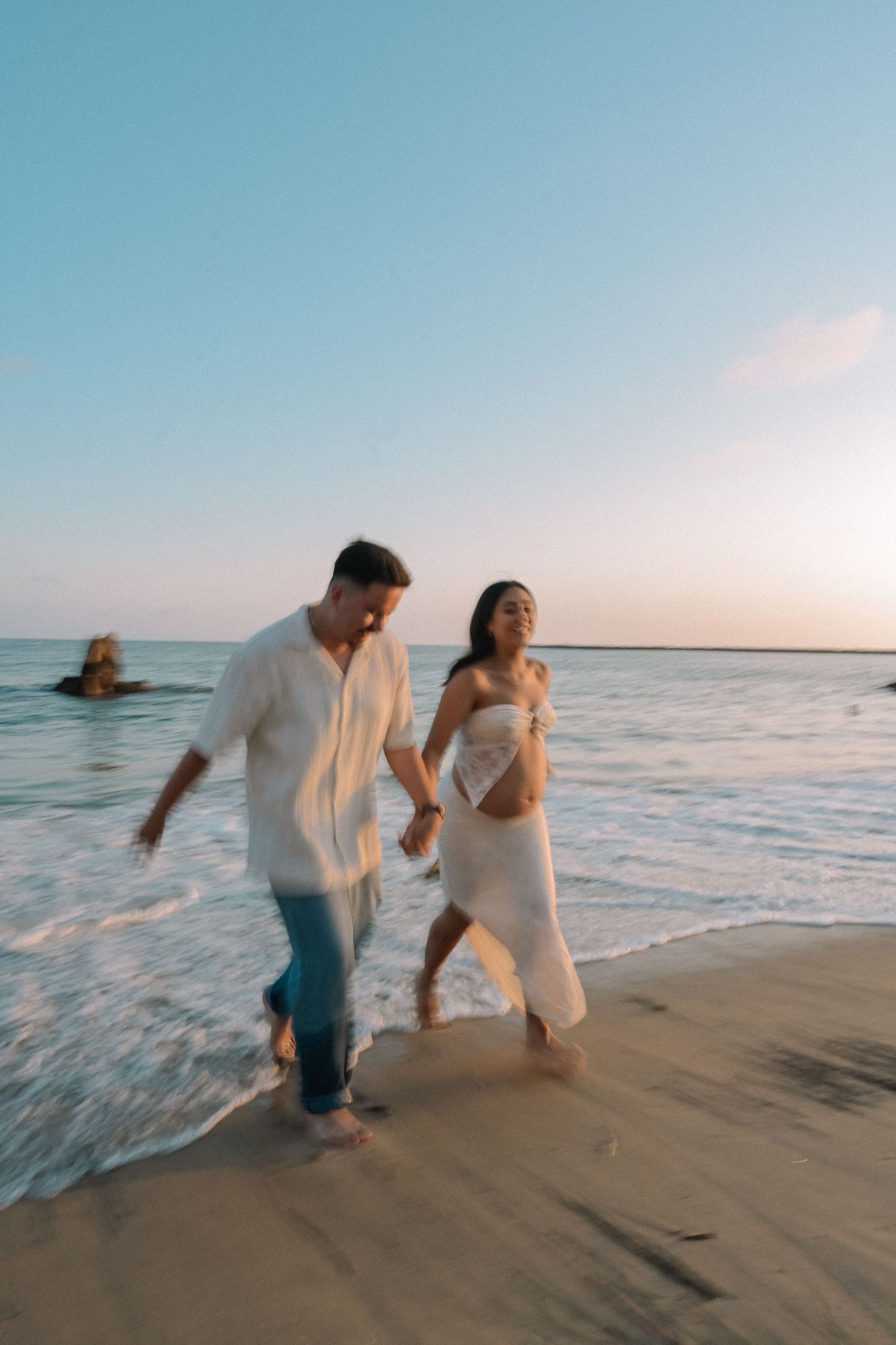 A couple in white attire shares romantic moments walking along a beach at sunset as waves wash ashore.