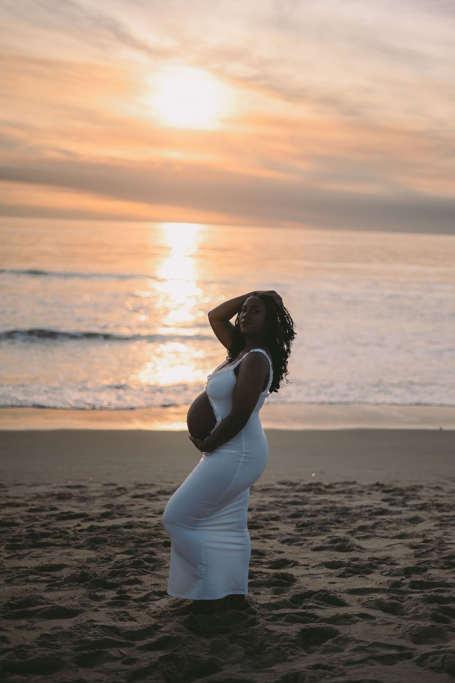 A silhouette of a pregnant woman in a white dress standing on a beach during golden hour sunset.