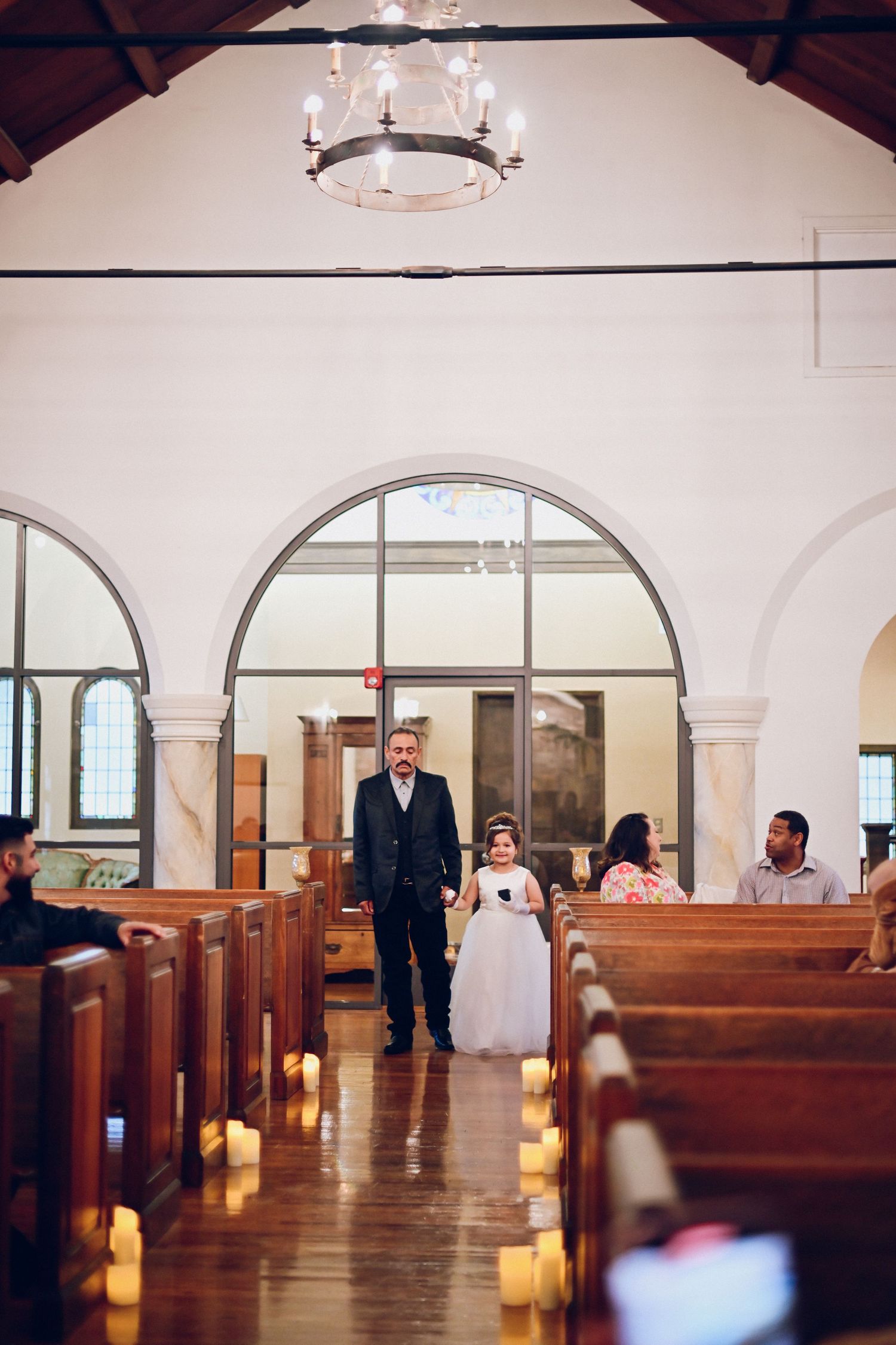 A procession walks down a church aisle lined with candles beneath an elegant chandelier.